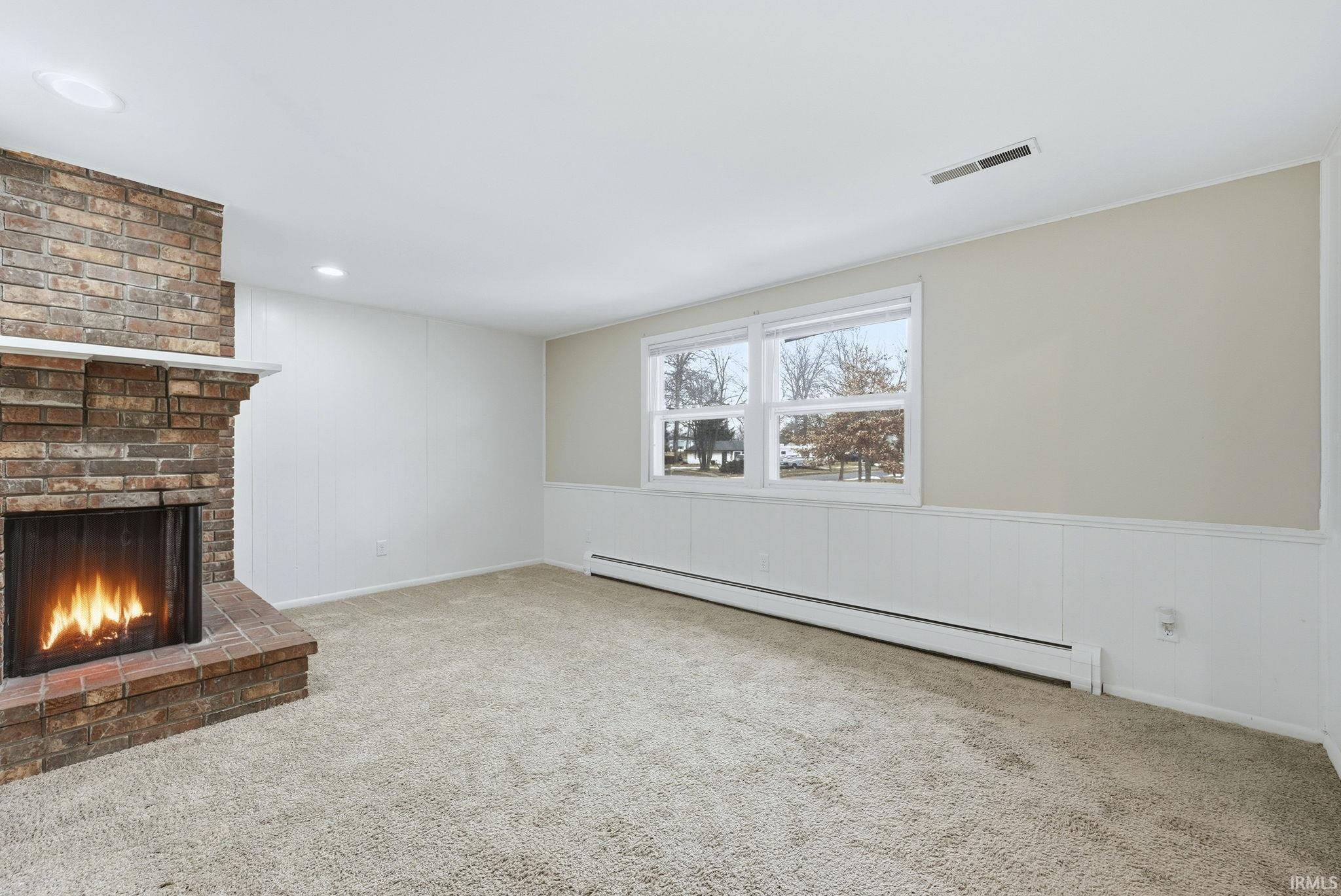 Unfurnished living room with a baseboard heating unit, light colored carpet, a brick fireplace, a wainscoted wall, and a decorative wall
