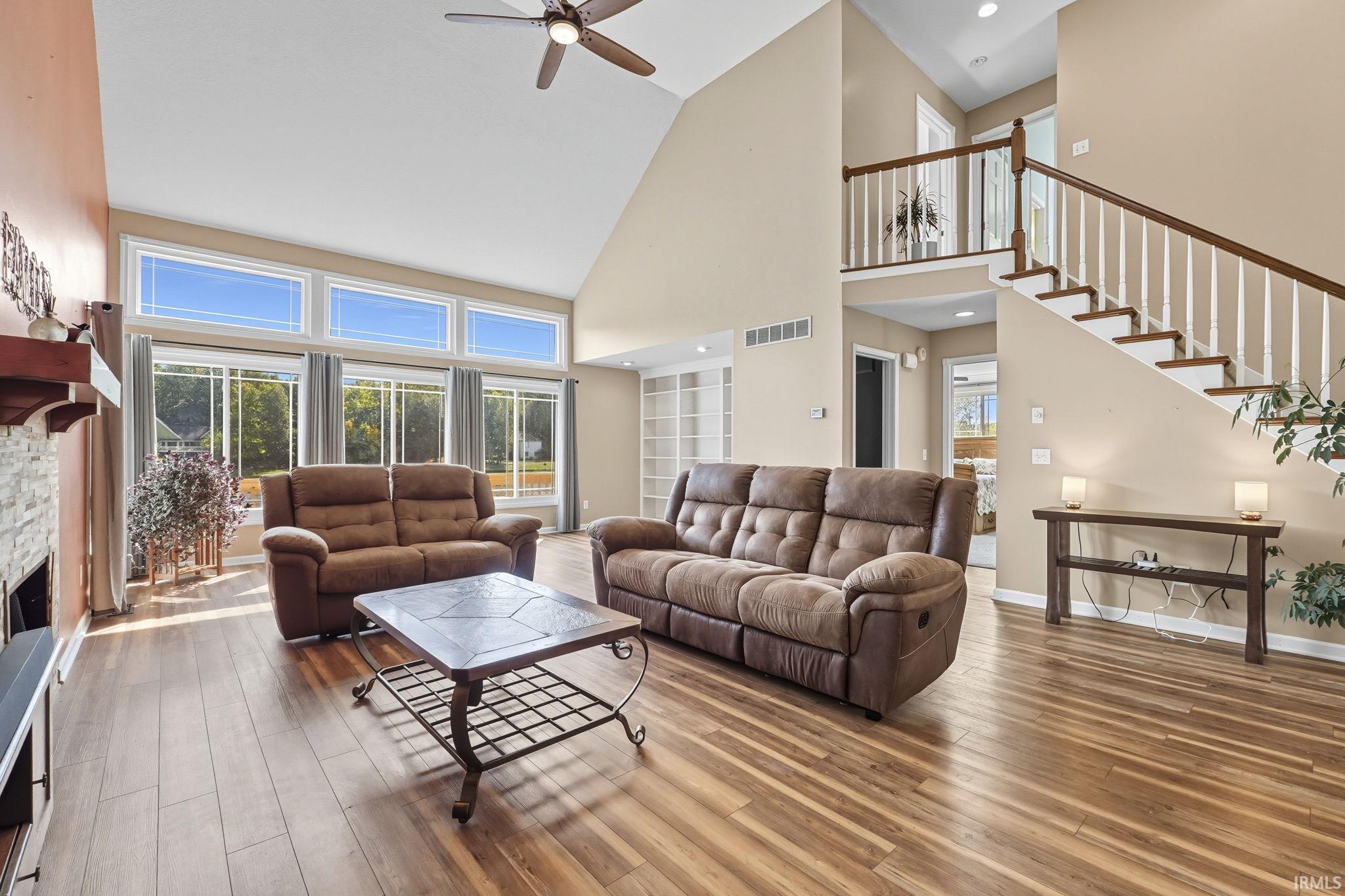 Living area with a ceiling fan, a stone fireplace, wood finished floors, and a high ceiling