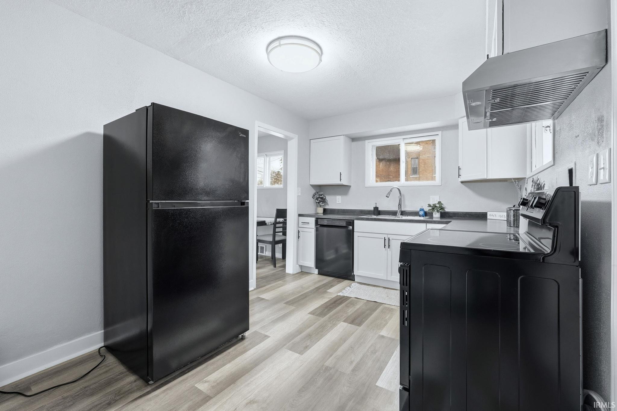 Kitchen with black appliances, white cabinetry, exhaust hood, dark countertops, and light wood-style flooring