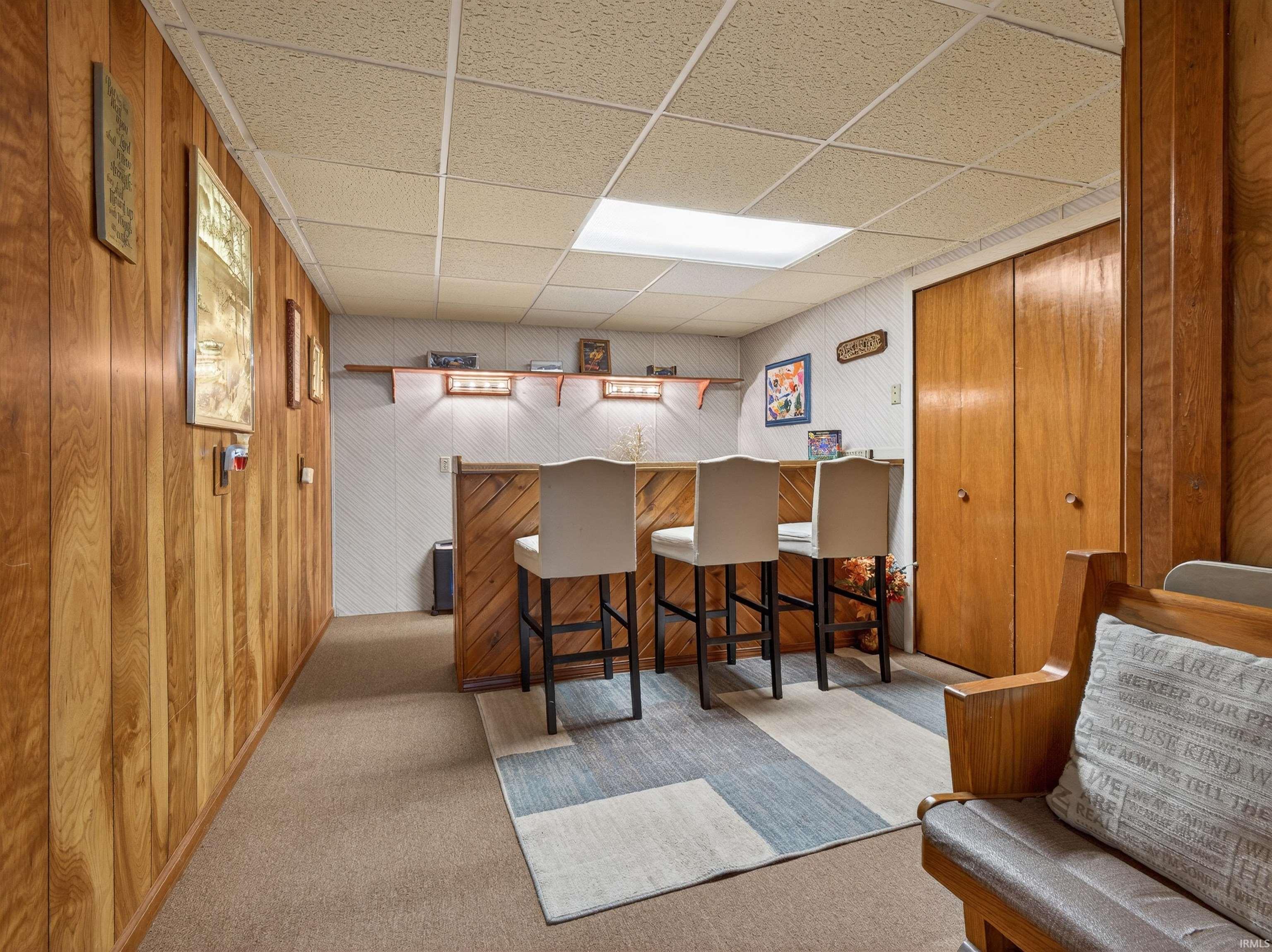 Indoor dry bar with a paneled ceiling, light colored carpet, and wooden walls