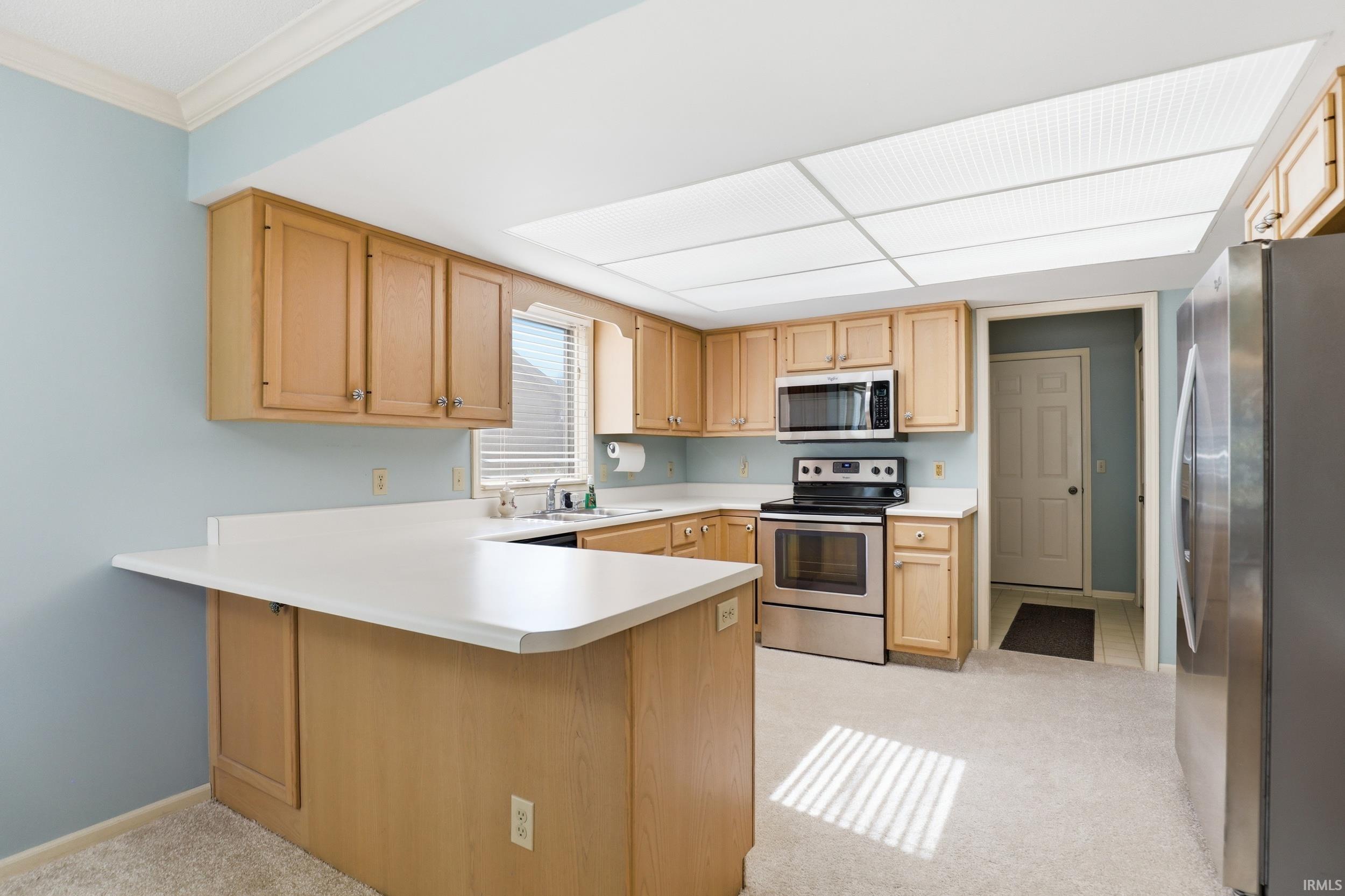 Kitchen with a peninsula, stainless steel appliances, light countertops, light colored carpet, and ornamental molding