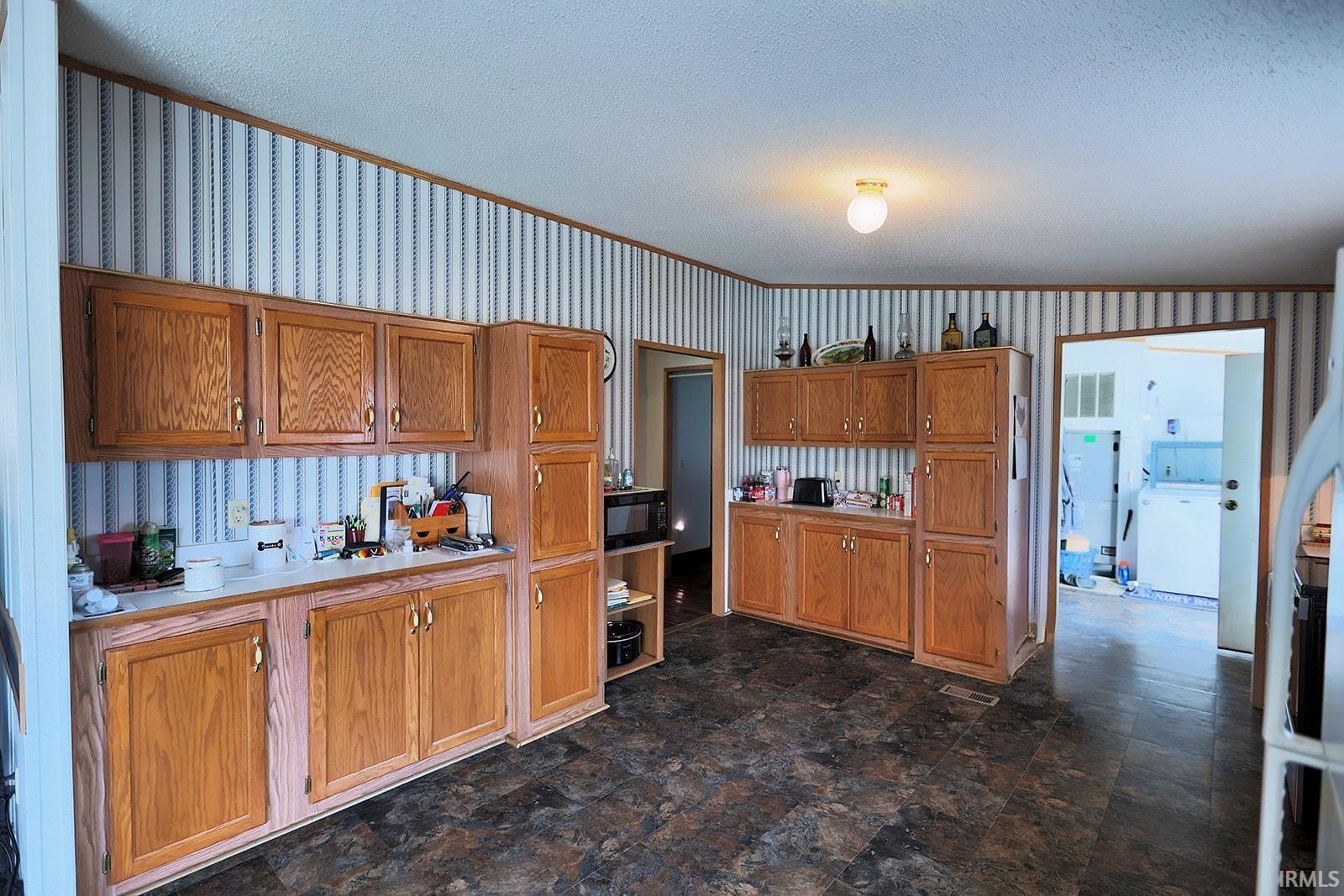 Kitchen featuring wood finish cabinetry, a textured ceiling, wallpapered walls, light countertops, and stone finish floors