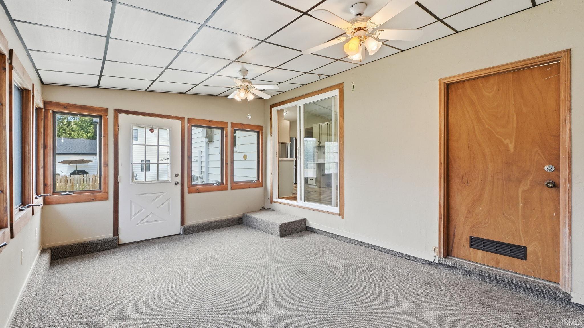 Enclosed sunroom featuring light gray carpet, multiple windows with wood trim, a white paneled door with windowpanes, a natural wood door, and two ceiling fans