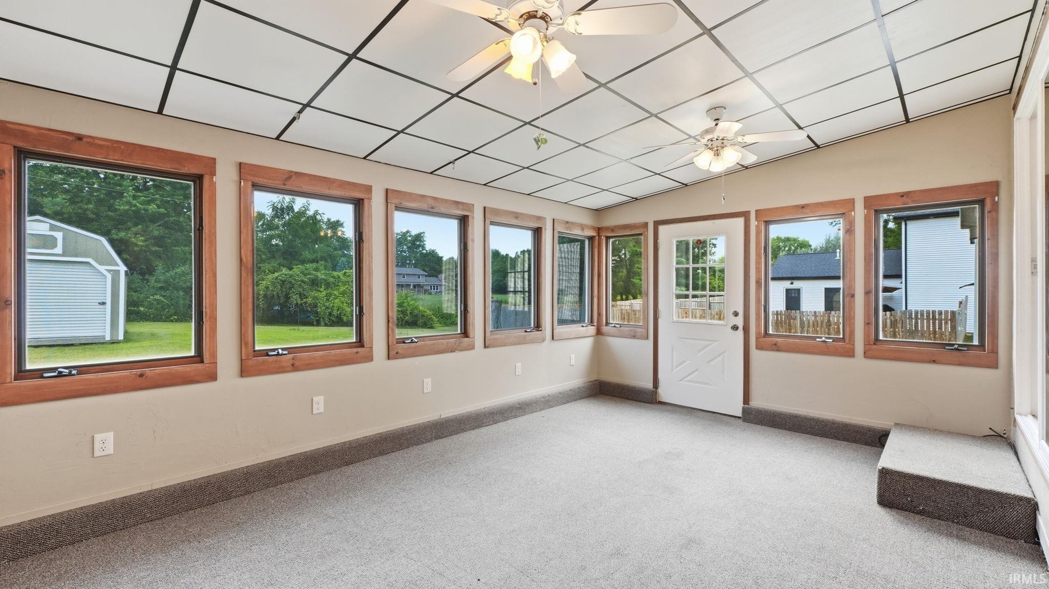 Sunroom featuring multiple windows with natural wood trim, wall-to-wall carpeting, a drop ceiling with integrated lighting and ceiling fans, and a glass-paneled exterior door