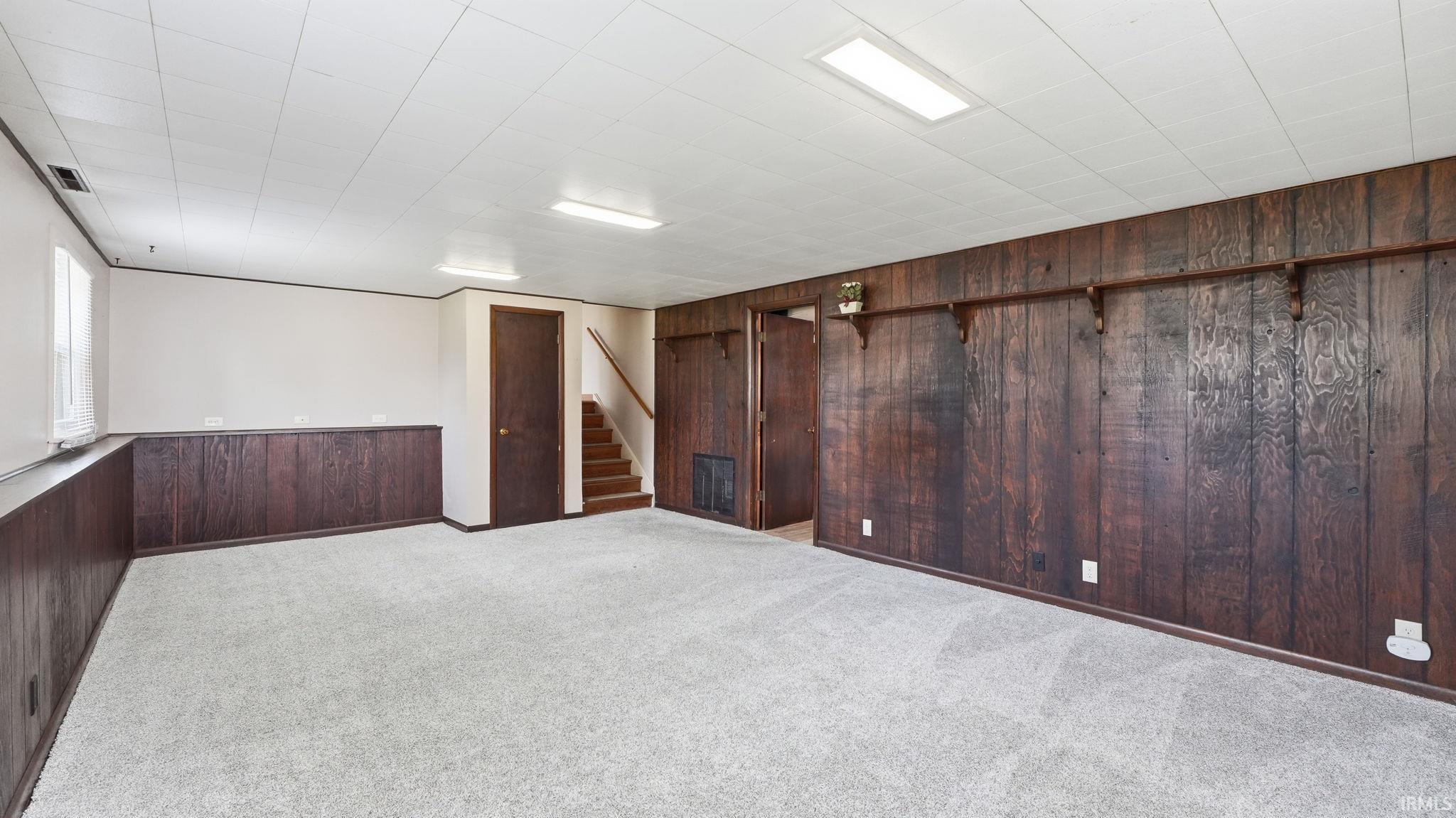 Spacious finished room featuring light gray carpeting, dark wood paneling, a white paneled ceiling with flush-mounted lighting, and a window providing natural illumination