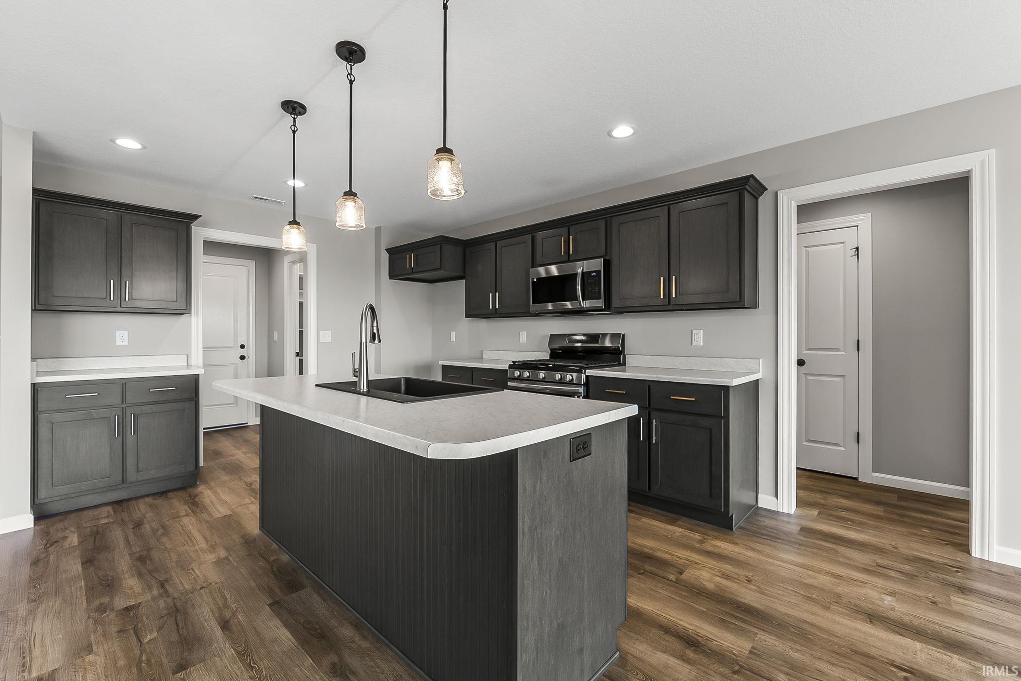 Kitchen featuring light countertops, stainless steel appliances, a center island with sink, dark wood-style floors, and hanging light fixtures