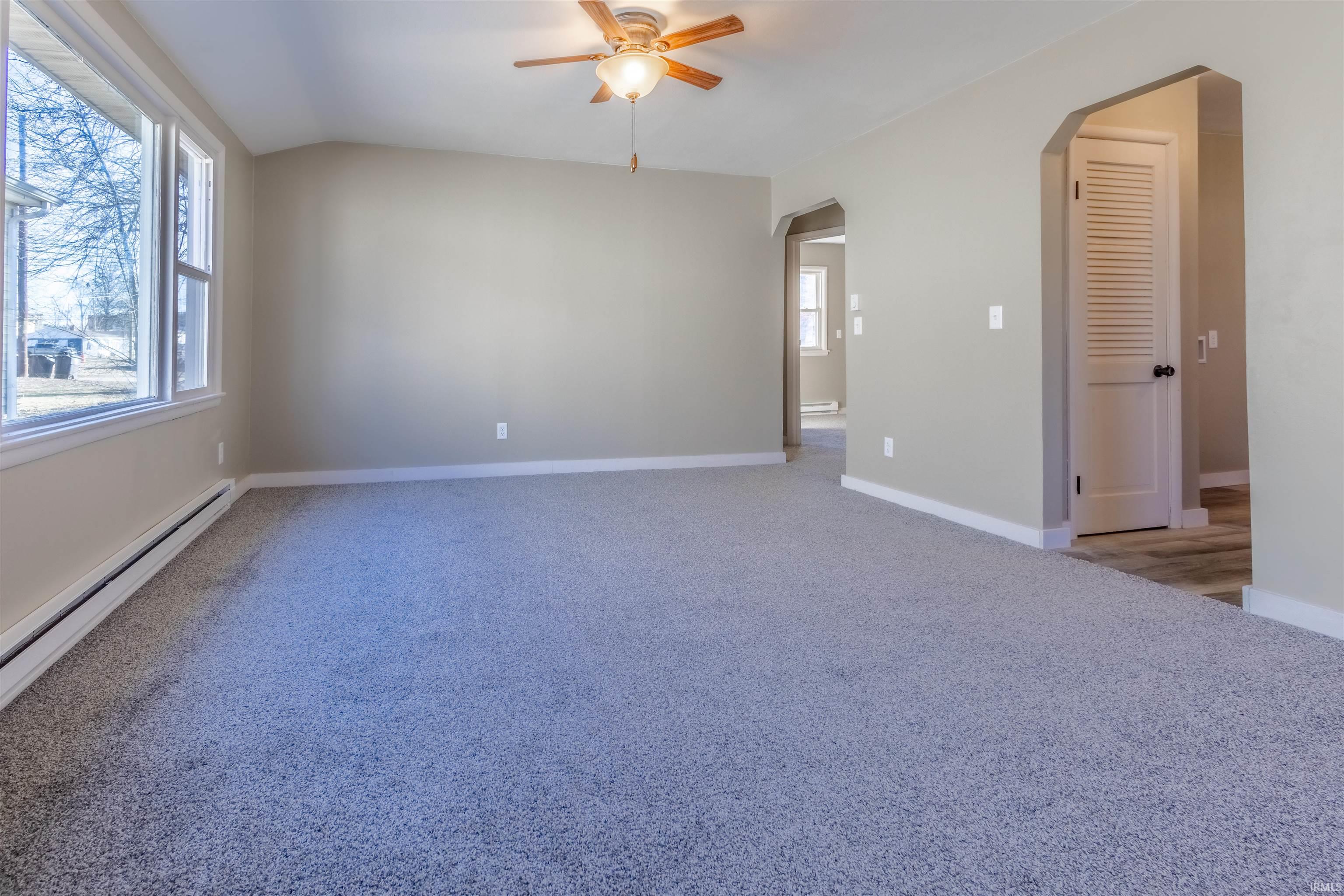 Unfurnished room featuring light colored carpet, ceiling fan, arched walkways, a baseboard radiator, and vaulted ceiling