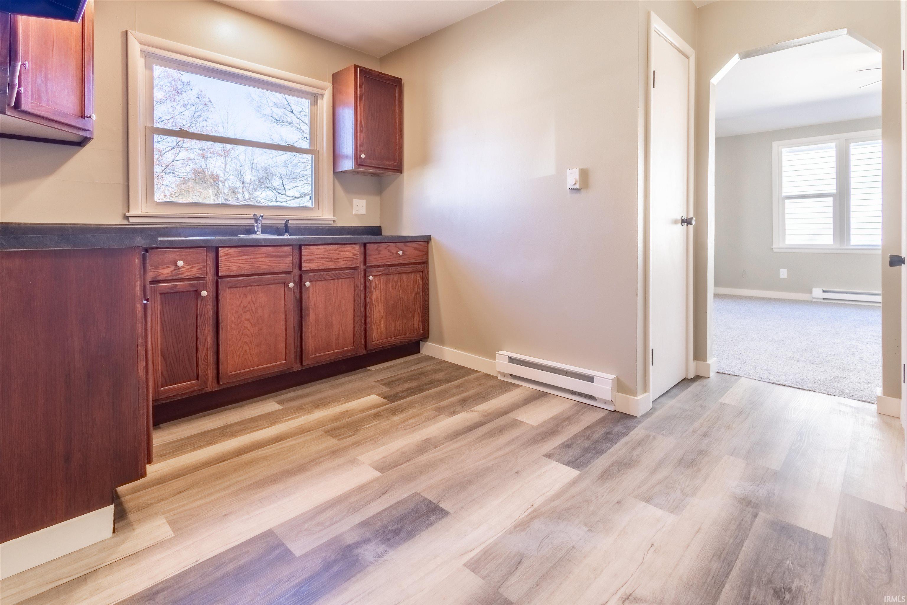 Kitchen with arched walkways, dark countertops, baseboard heating, light wood-style floors, and plenty of natural light