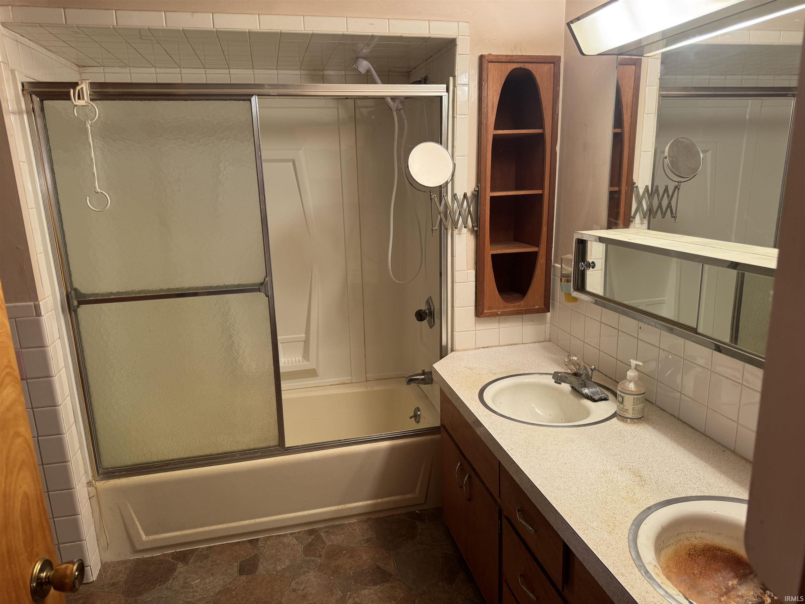 Bathroom featuring double vanity, enclosed tub / shower combo, stone finish flooring, and backsplash
