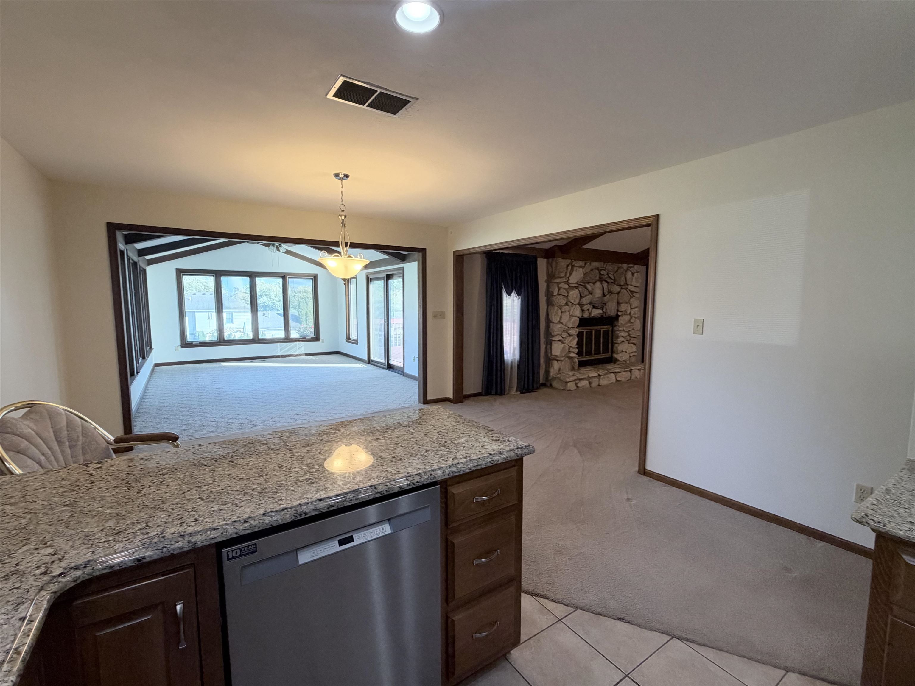 Kitchen featuring a stone-patterned countertop, stainless steel dishwasher, and wooden cabinetry, accompanied by a stone masonry fireplace and a multi-windowed sunroom with vaulted ceiling beams