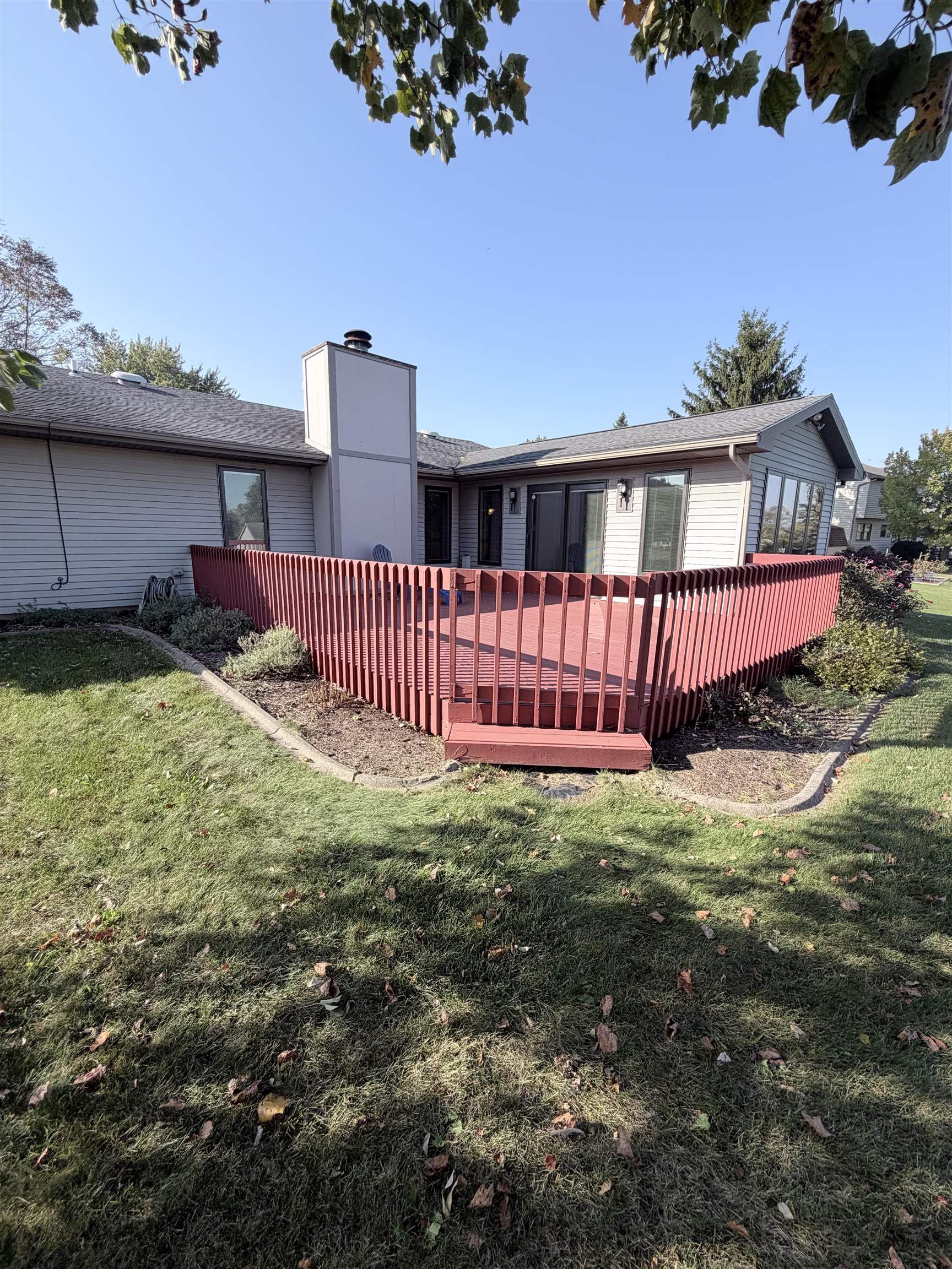 Elevated rear deck with horizontal balusters, exterior masonry chimney, and sliding glass door access, set against a manicured lawn with defined stone landscape edging