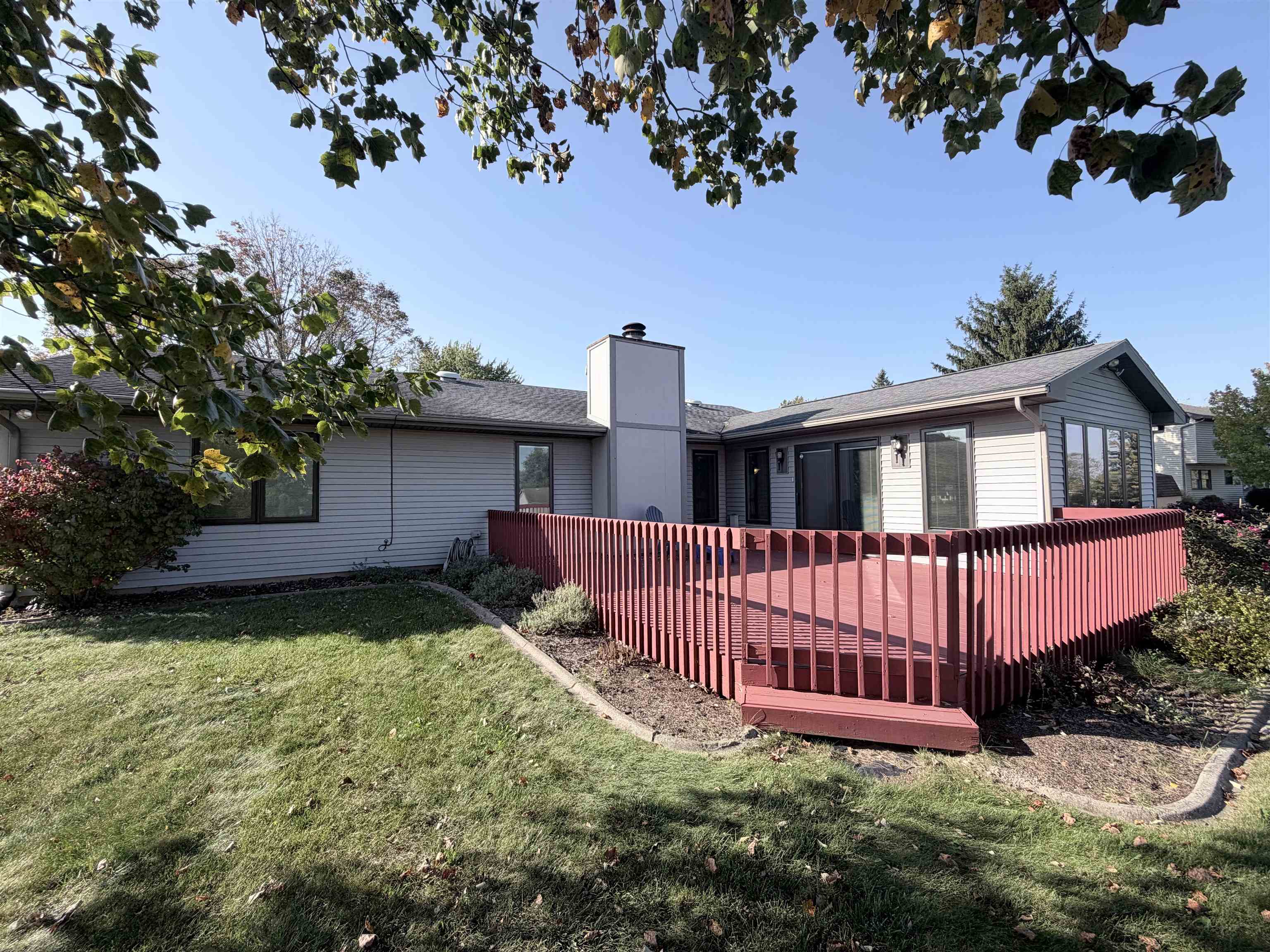Exterior of the property featuring a raised wood deck with perimeter railing, central chimney, vinyl siding, and landscaped lawn with stone edging