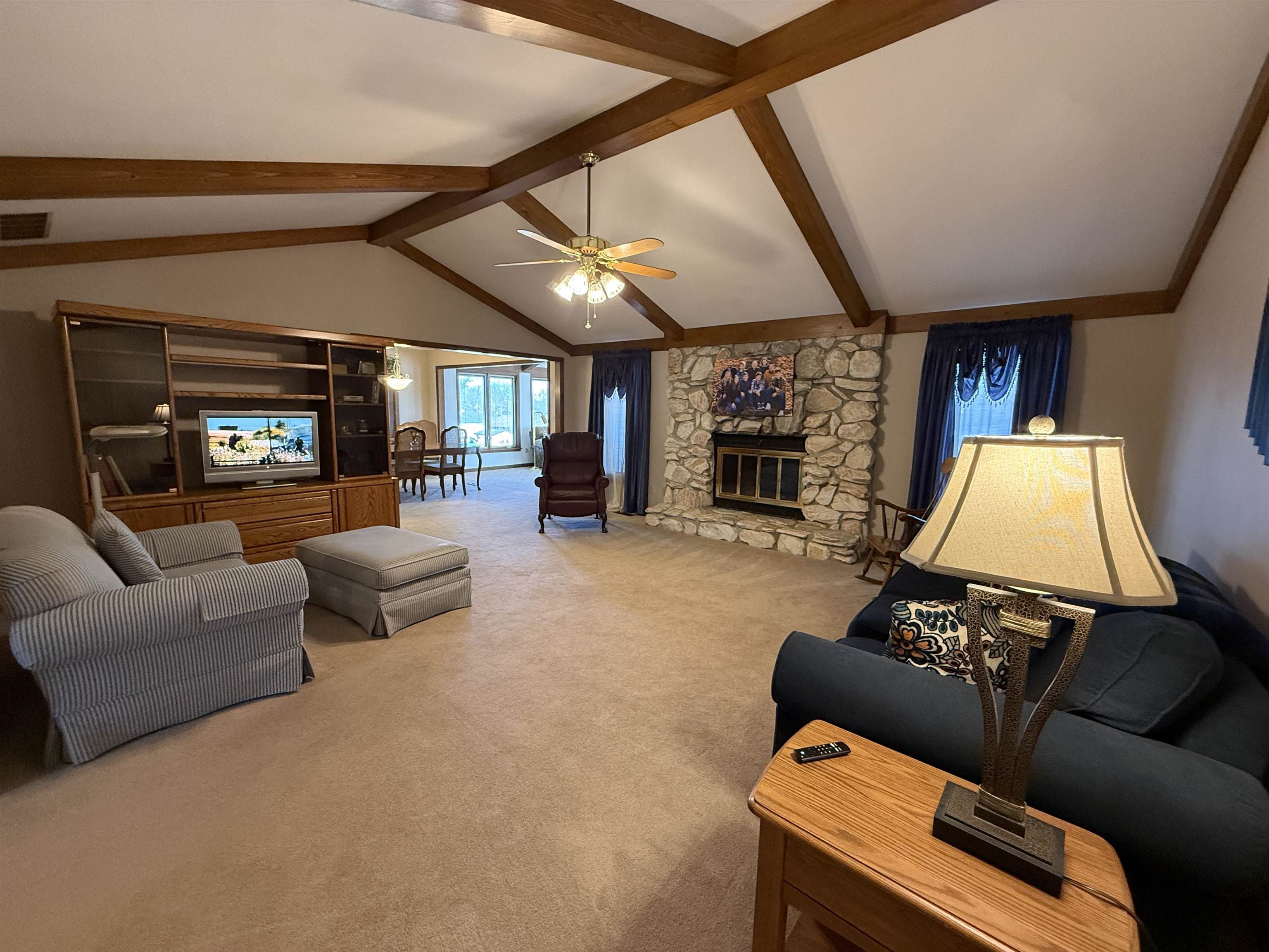Carpeted living room featuring a stone fireplace, ceiling fan, and beam ceiling