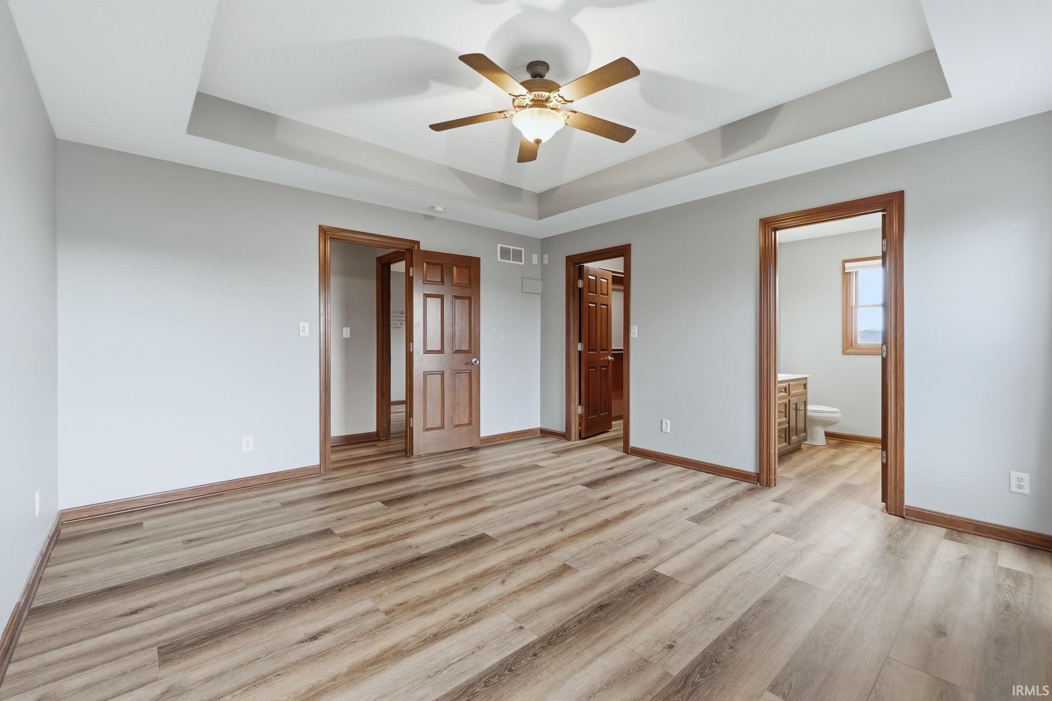 Unfurnished bedroom with a spacious closet, a raised ceiling, light wood-type flooring, ceiling fan, and ensuite bath