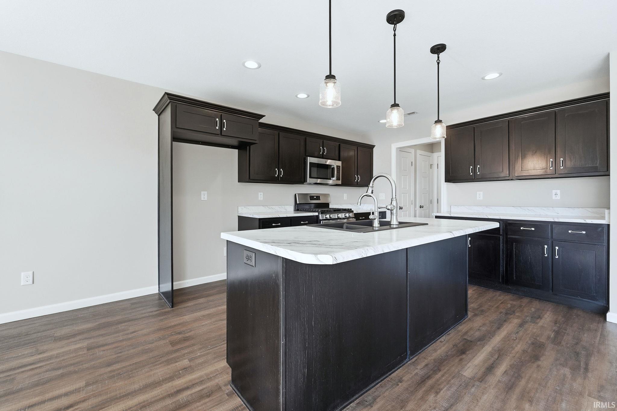 Kitchen featuring light countertops, stainless steel appliances, hanging light fixtures, dark wood-style floors, and a center island with sink