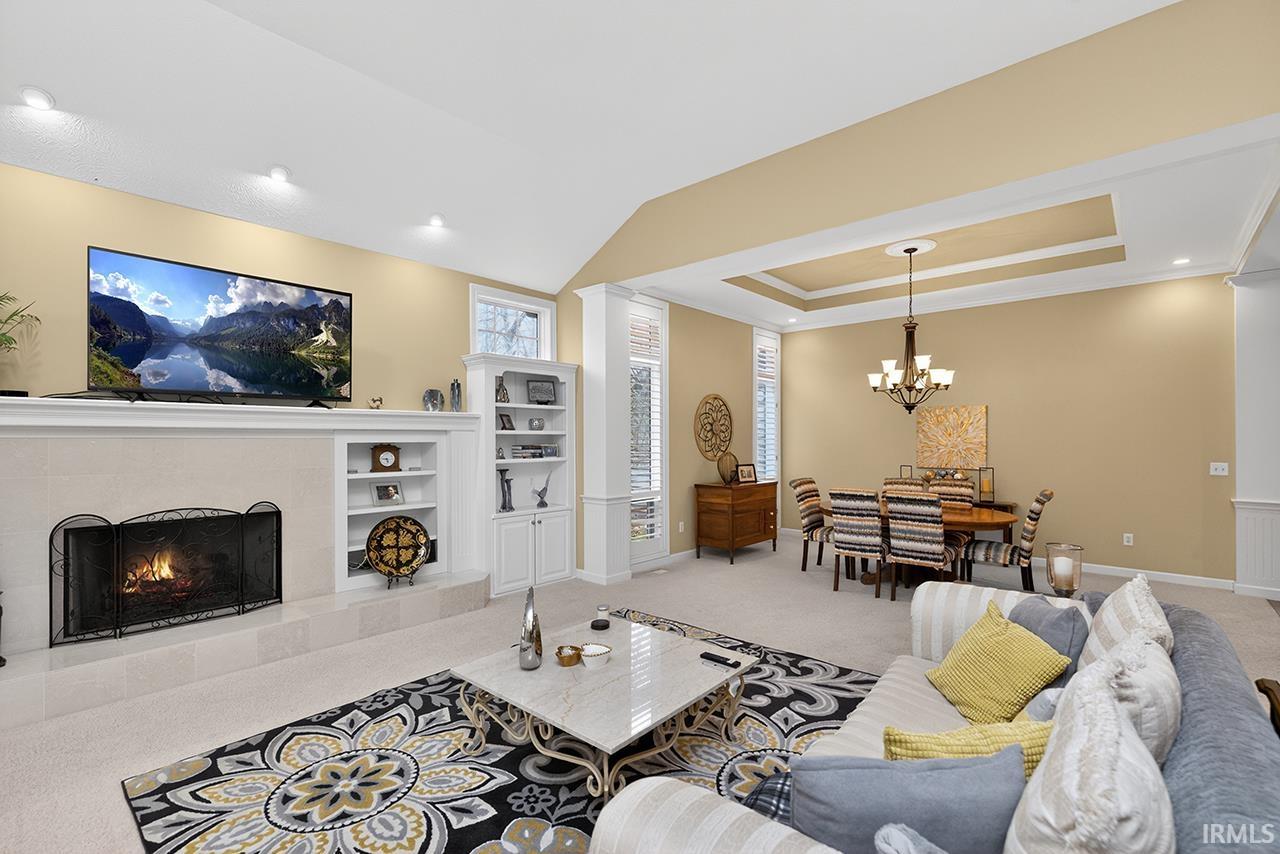 Living room with light carpet, a tiled fireplace, a tray ceiling, built in features, and decorative columns
