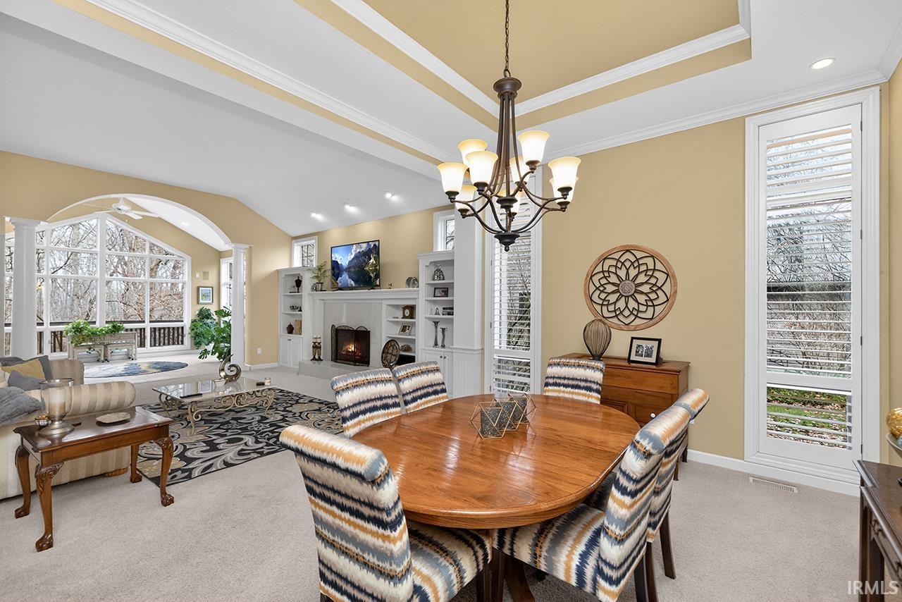 Dining area featuring a lit fireplace, light colored carpet, hanging lights, ornamental molding, and a tray ceiling
