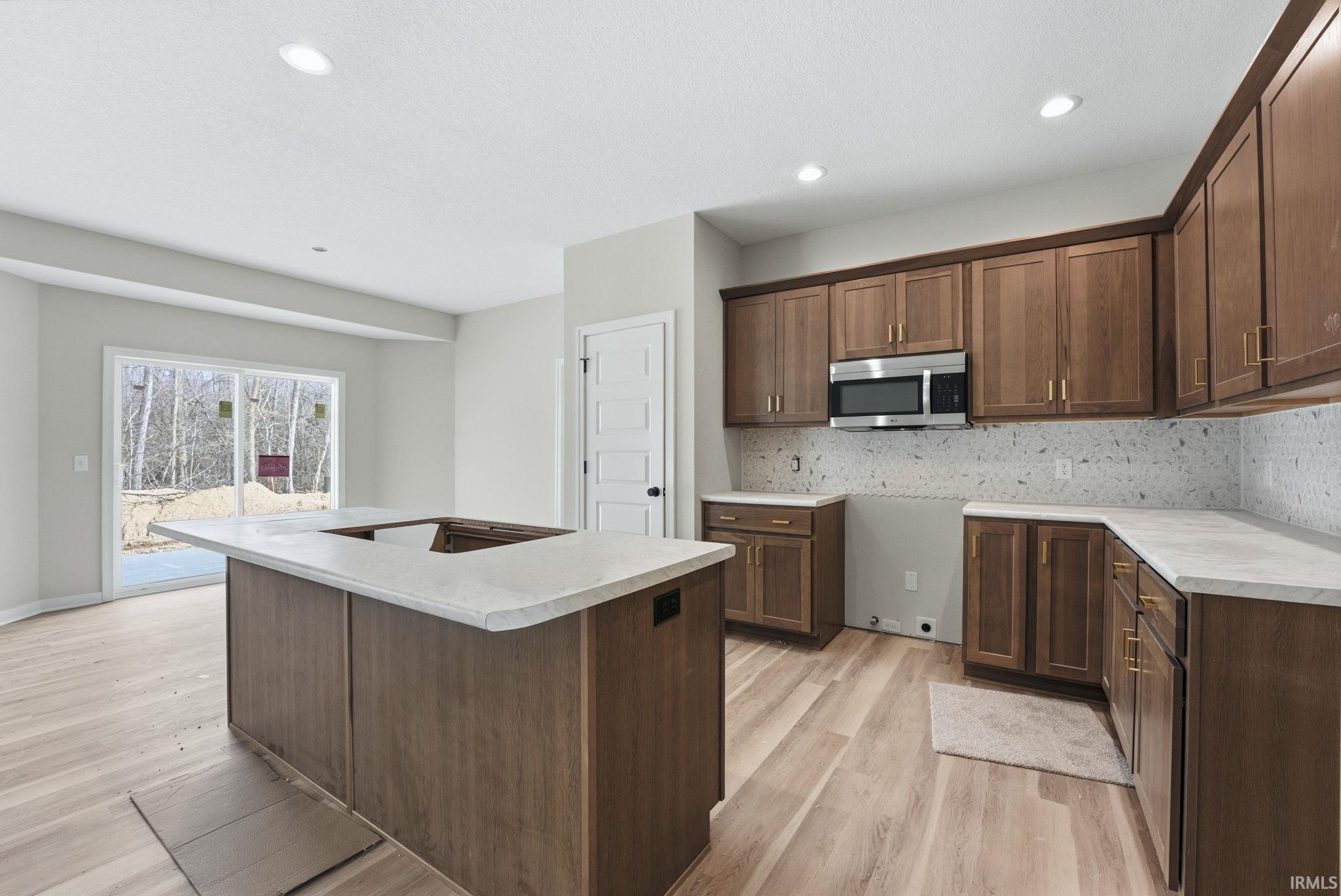 Kitchen with light countertops, light wood finished floors, stainless steel microwave, a kitchen island with sink, and backsplash