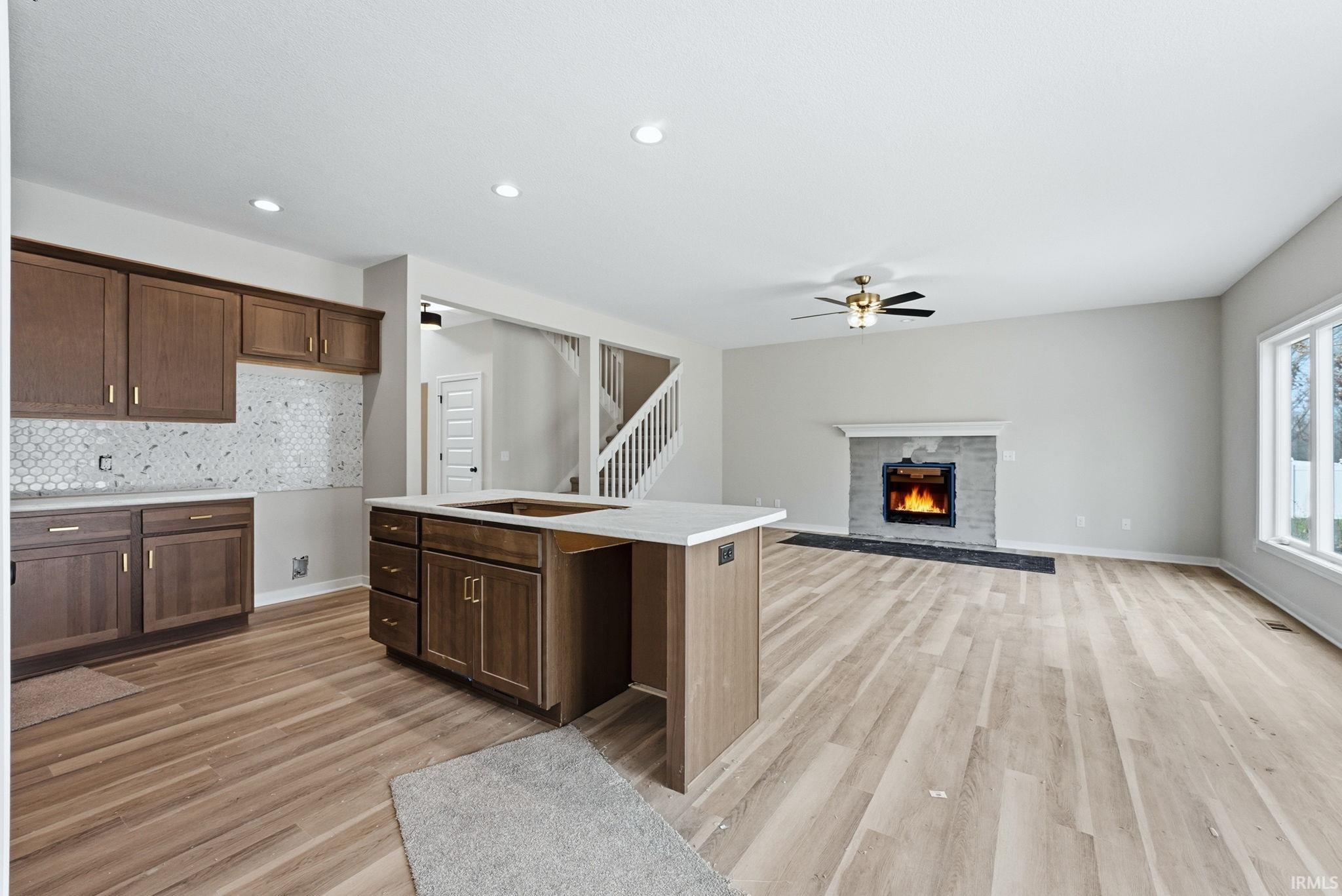 Kitchen featuring light wood-style flooring, a kitchen island with sink, a warm lit fireplace, backsplash, and recessed lighting