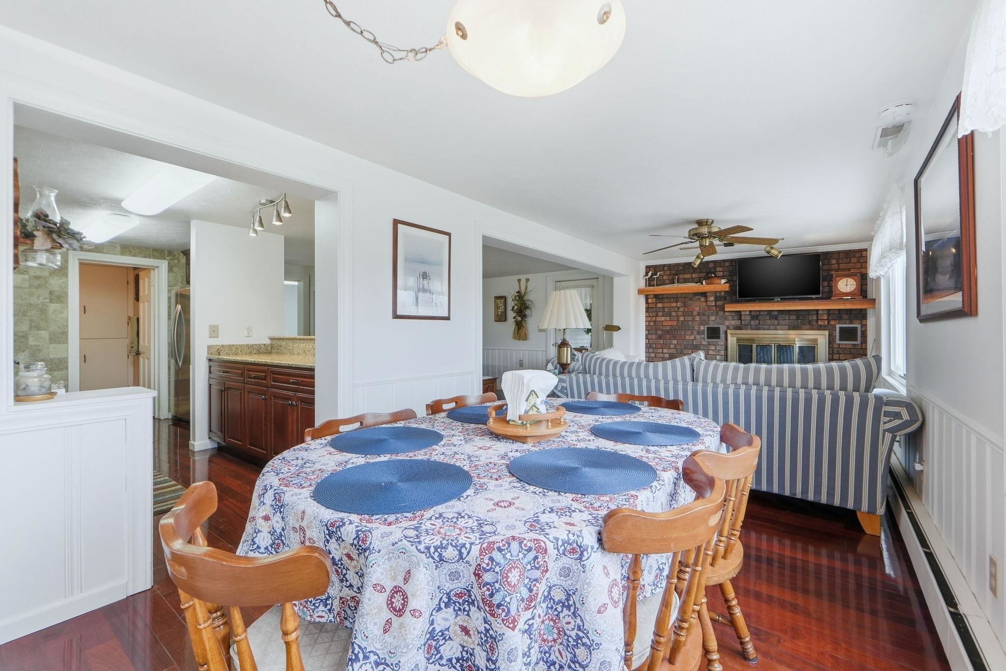 Dining room with baseboard heating, dark wood-style flooring, a ceiling fan, a wainscoted wall, and a brick fireplace