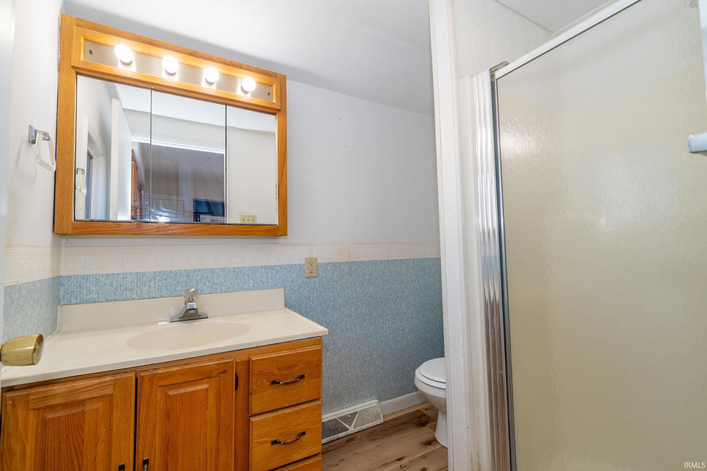 Full bathroom featuring vanity, wallpapered walls, light wood-type flooring, a shower stall, and wainscoting