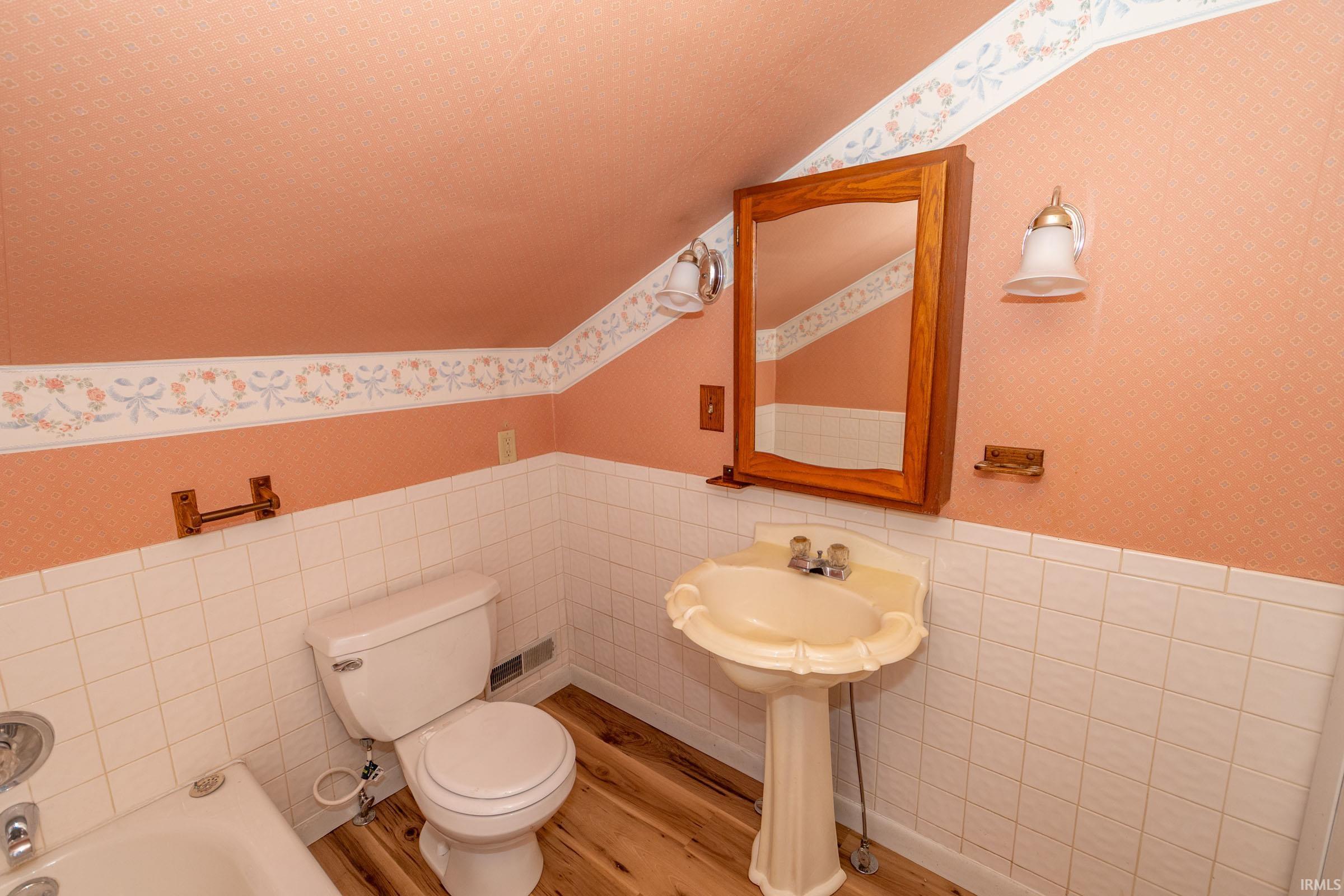 Bathroom featuring wallpapered walls, tile walls, a wainscoted wall, wood finished floors, and a bath
