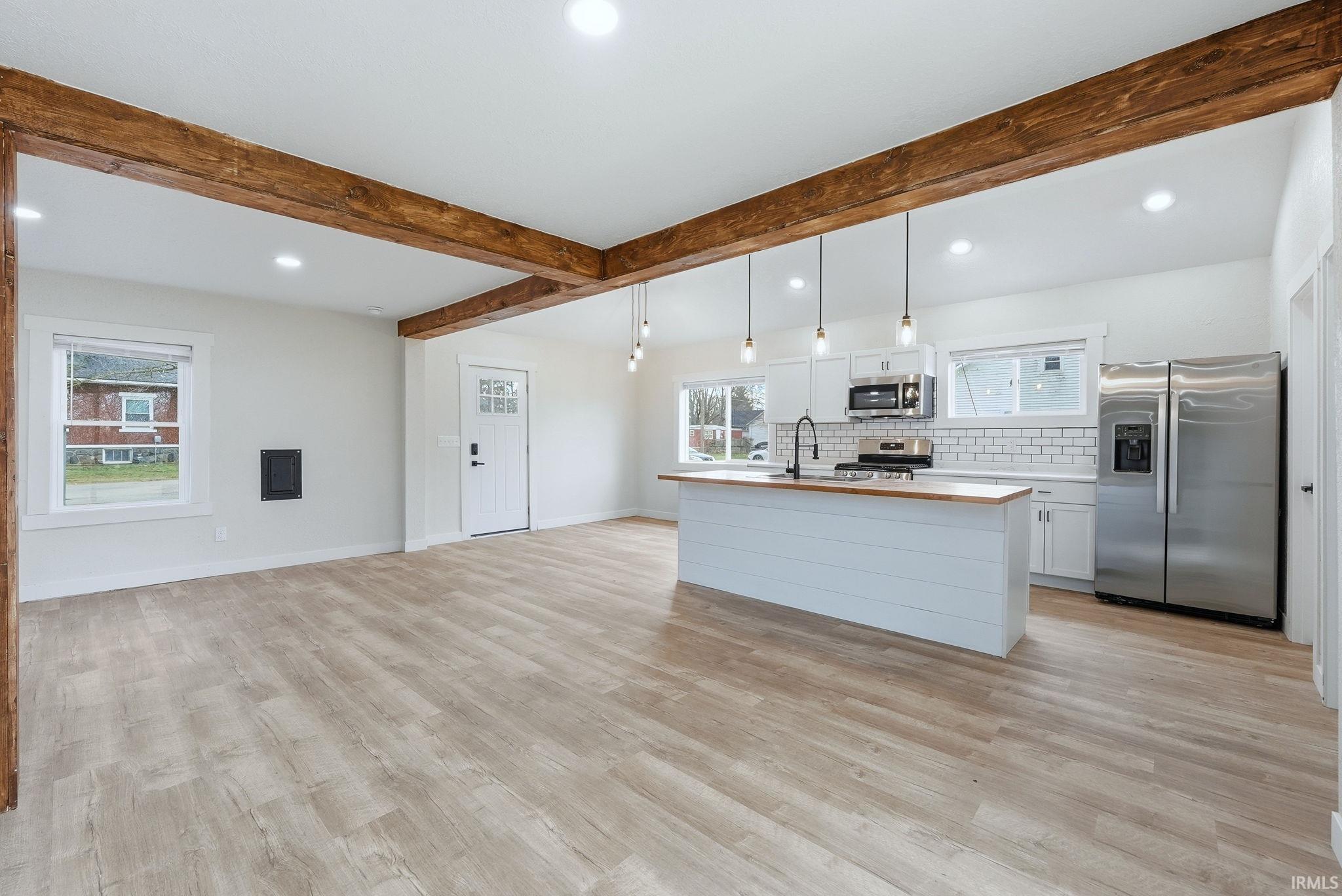 Kitchen featuring white cabinets, stainless steel appliances, an island with sink, backsplash, and beam ceiling