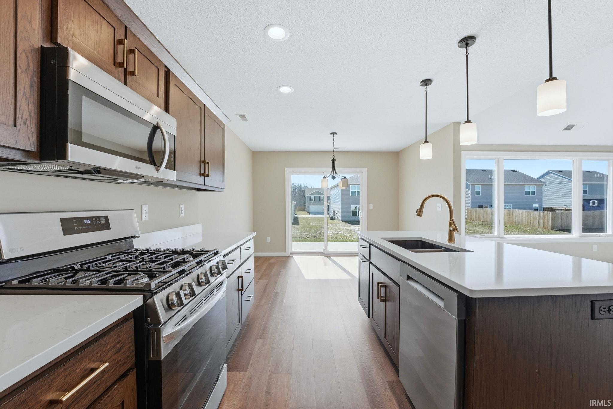 Kitchen with stainless steel appliances, hanging light fixtures, light wood finished floors, a kitchen island with sink, and light stone counters
