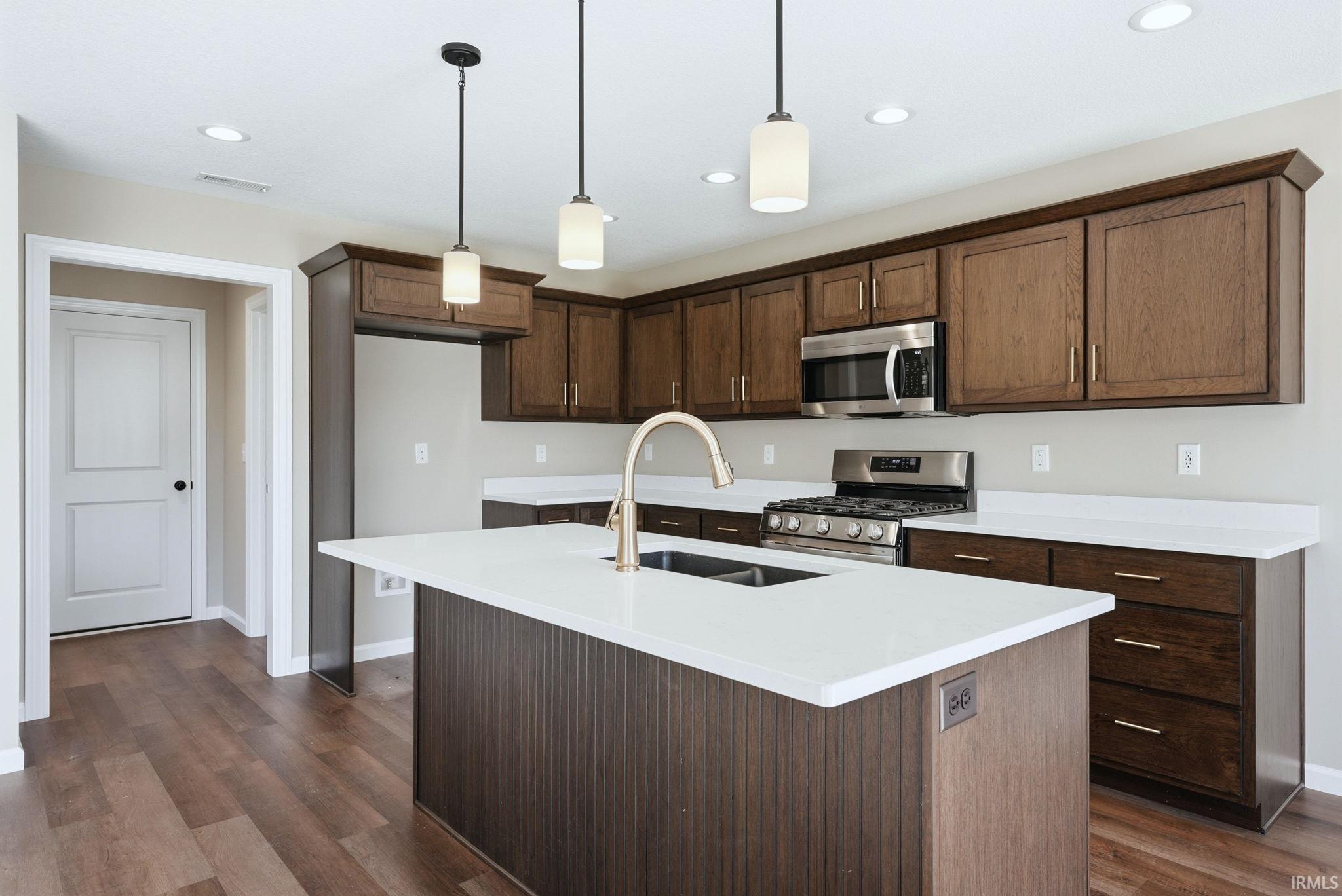 Kitchen with an island with sink, stainless steel appliances, decorative light fixtures, light stone countertops, and dark wood-style floors