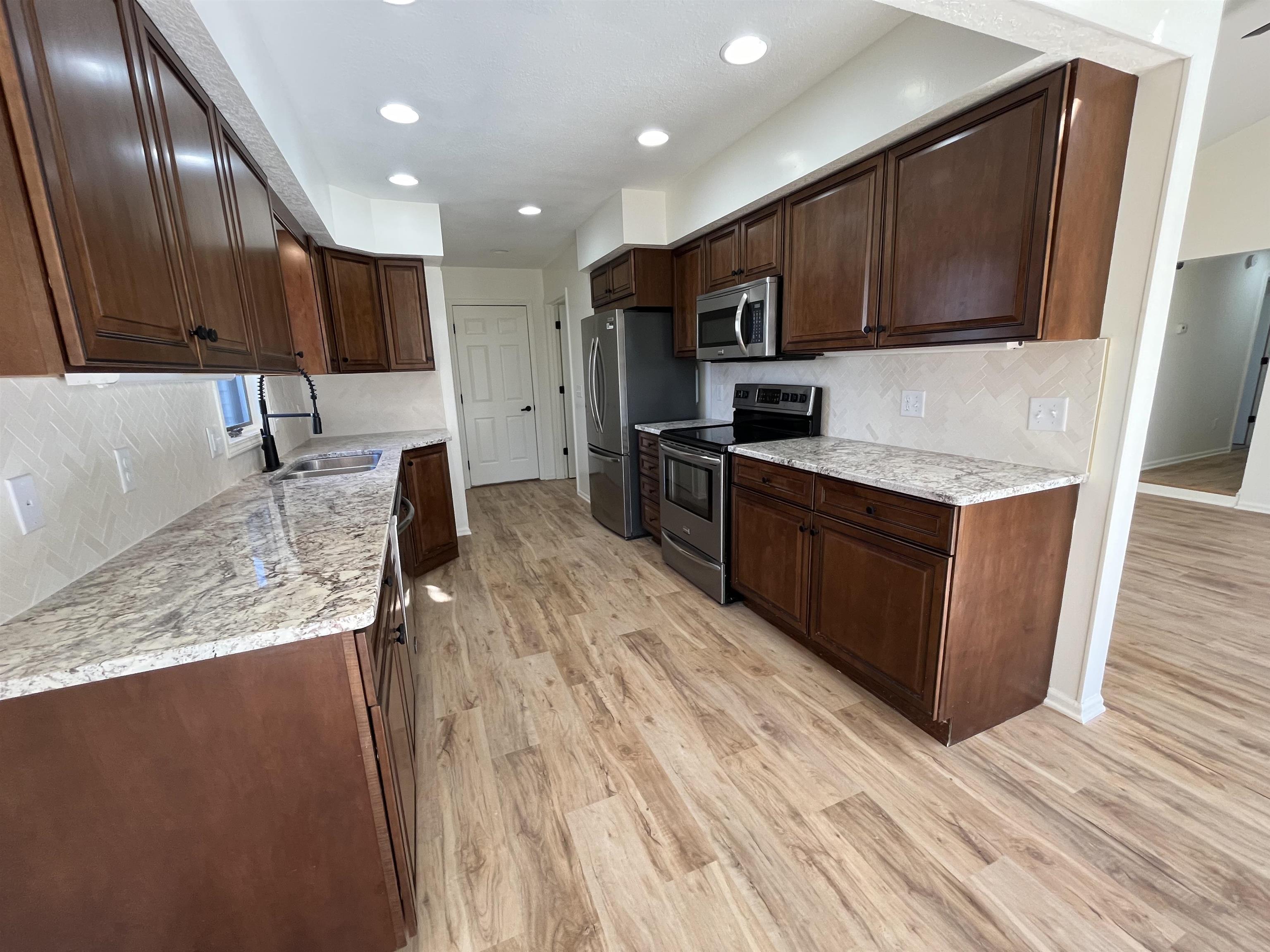 Kitchen with stainless steel appliances, backsplash, light wood-type flooring, light stone counters, and dark wood finish cabinets