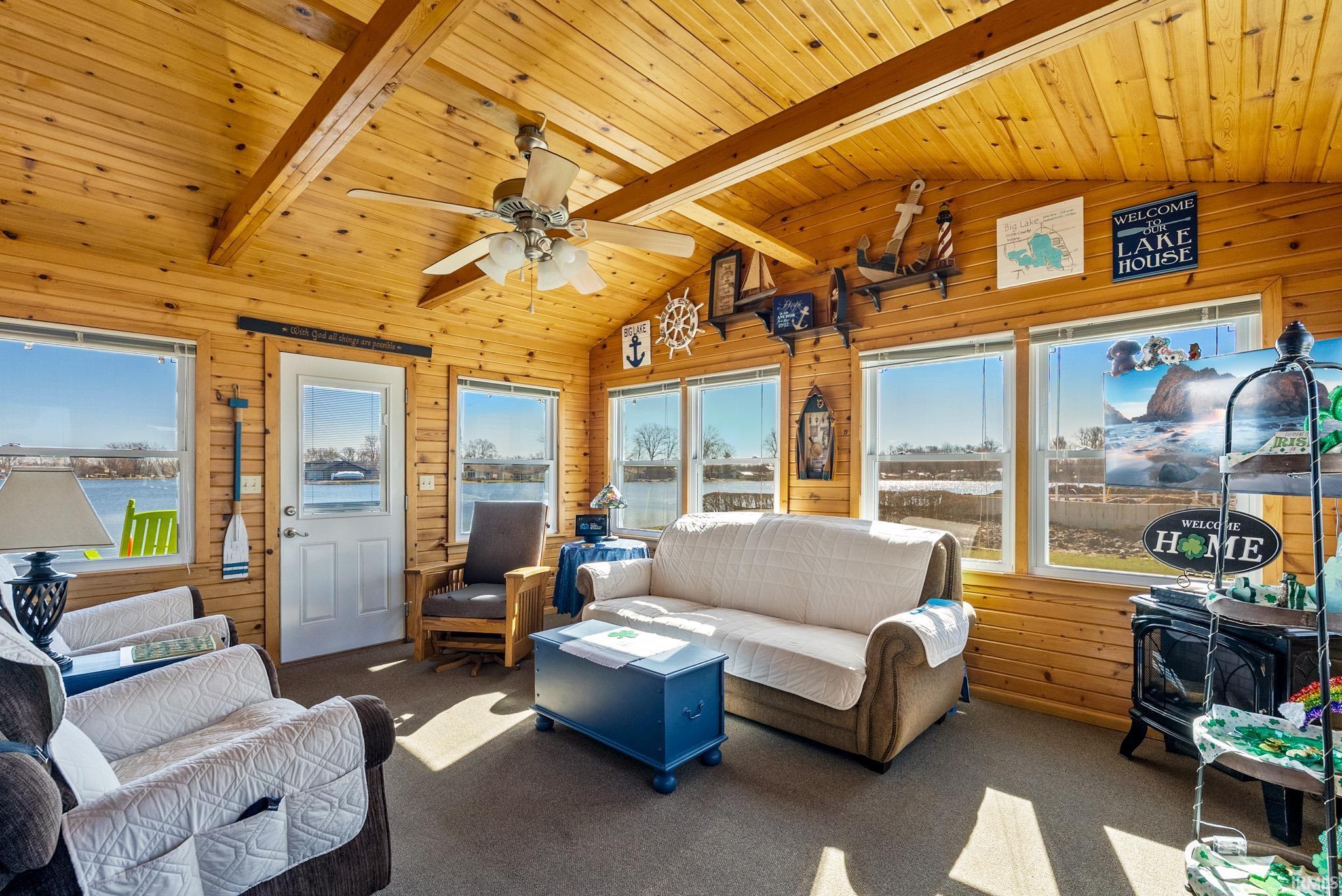Carpeted living area with wood walls, a wood stove, ceiling fan, and a wood ceiling with exposed beams