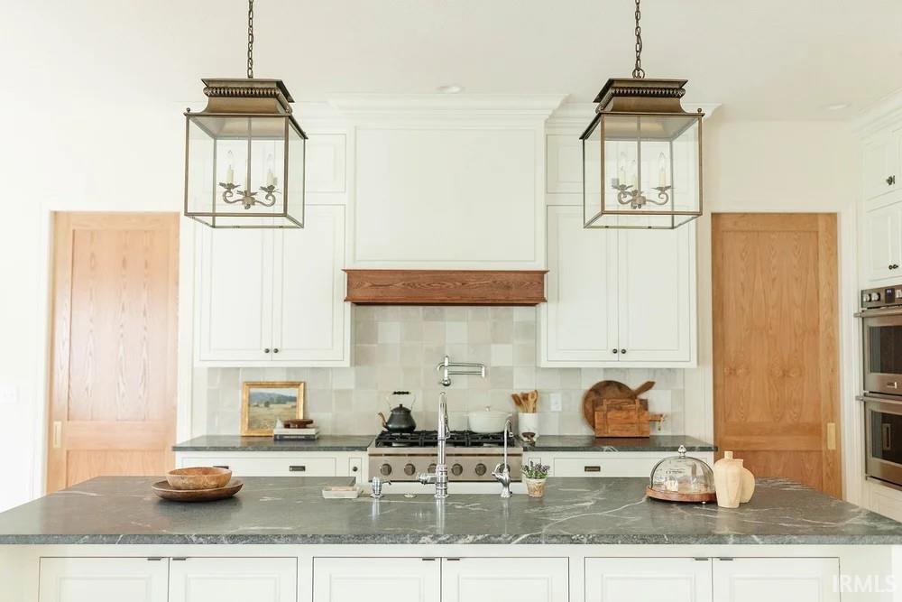 Kitchen featuring white cabinets, backsplash, dark stone countertops, suspended lighting, and double oven