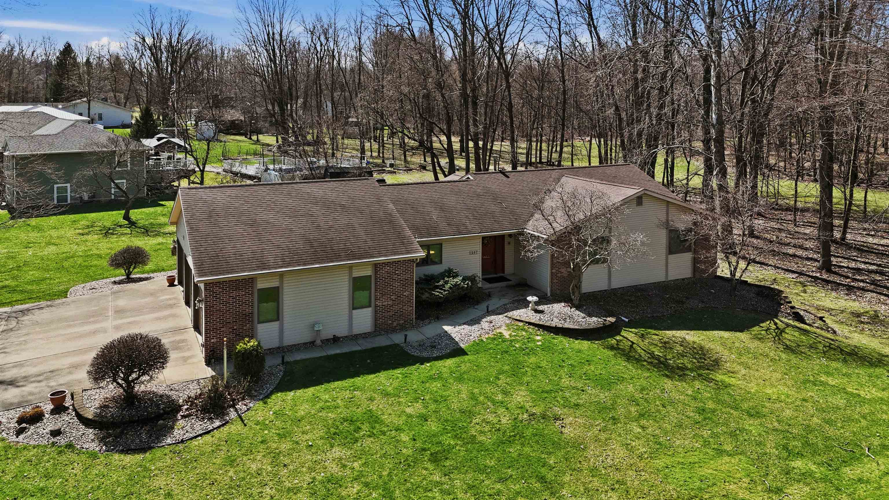 Single story home with a shingled roof, brick siding, a front yard, and view of wooded area
