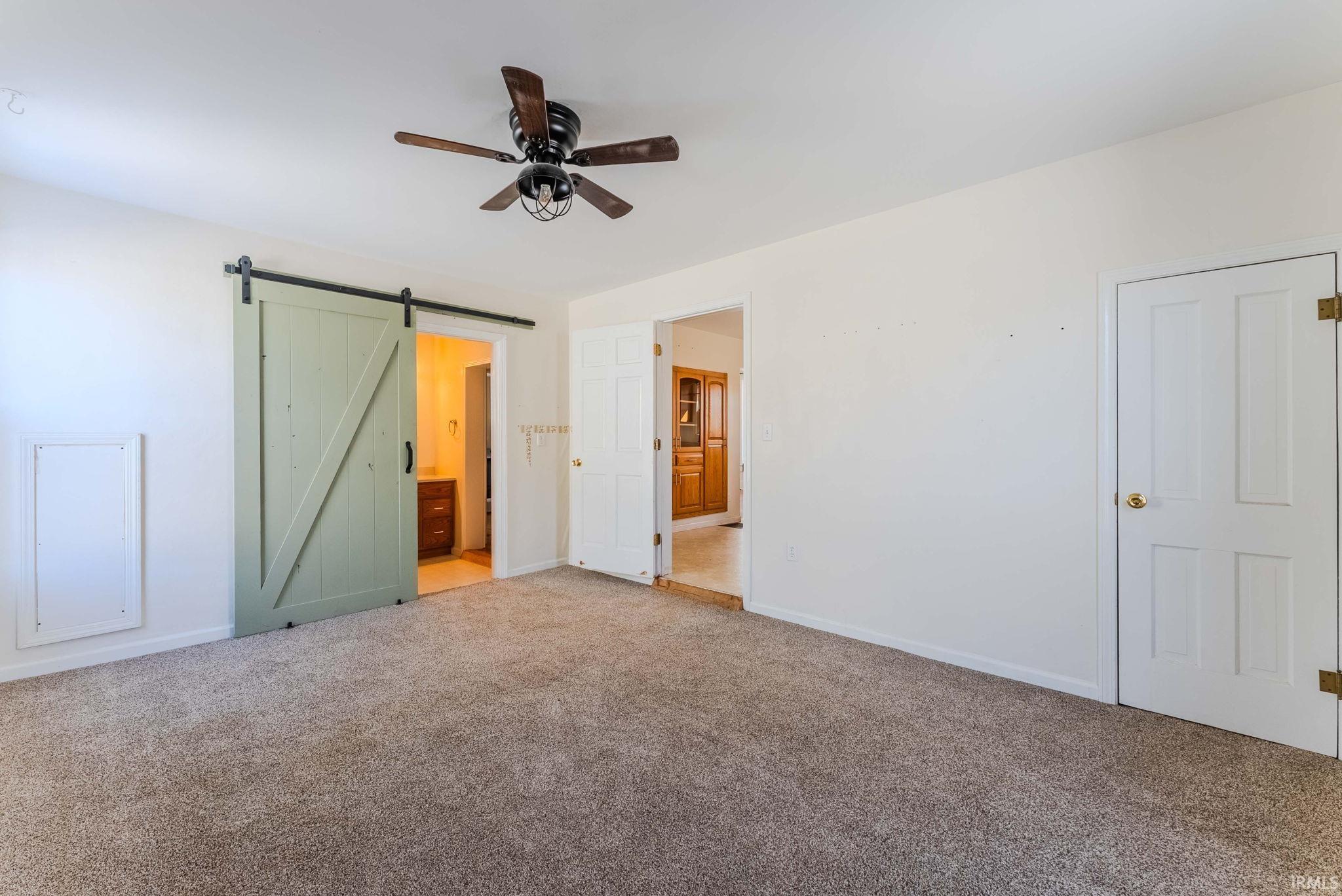 Unfurnished bedroom with a barn door, ensuite bath, carpet flooring, and a ceiling fan