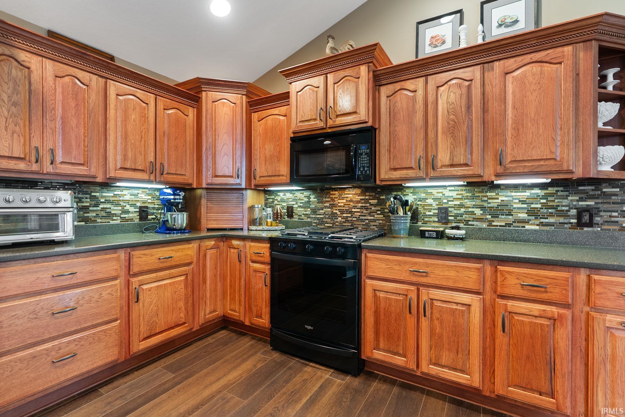 Kitchen with black appliances, wood finish cabinetry, dark wood-style floors, dark stone countertops, and lofted ceiling
