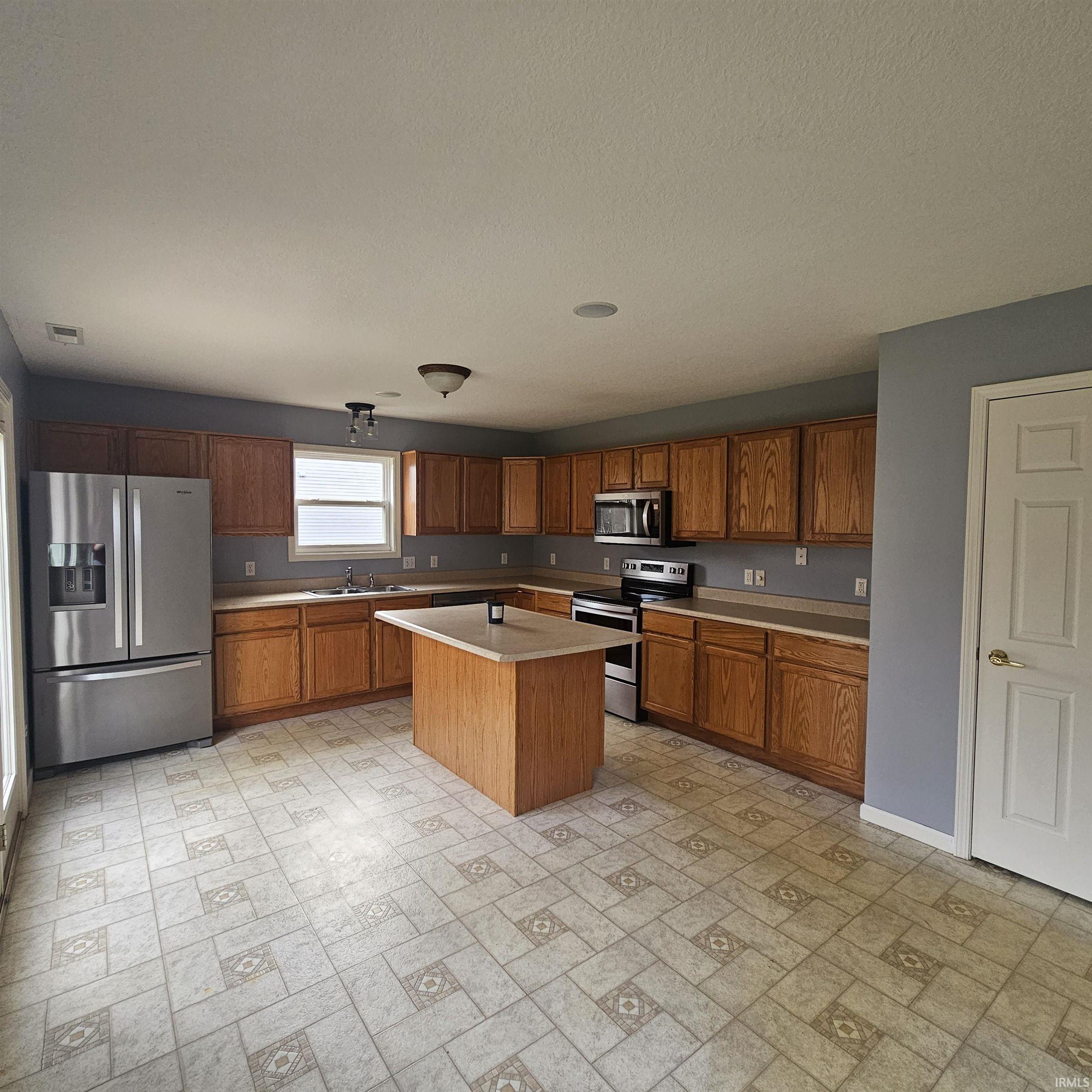 Kitchen featuring stainless steel appliances, wood finish cabinetry, light countertops, a center island, and brick patterned floors