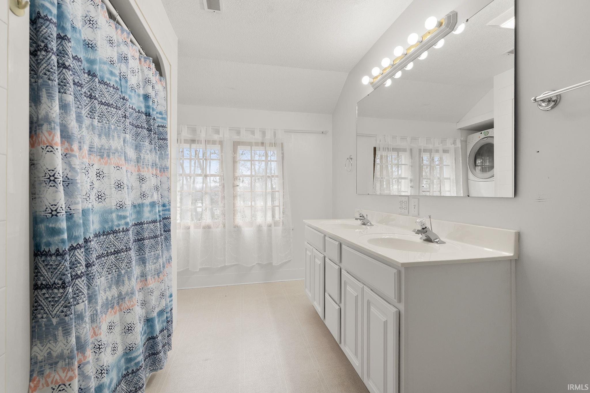 Full bathroom with washer / dryer, double vanity, vaulted ceiling, a shower with shower curtain, and light flooring