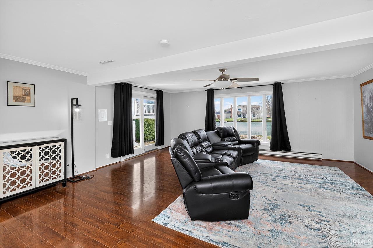 Living room with crown molding, dark wood-type flooring, a ceiling fan, a water view, and a baseboard radiator