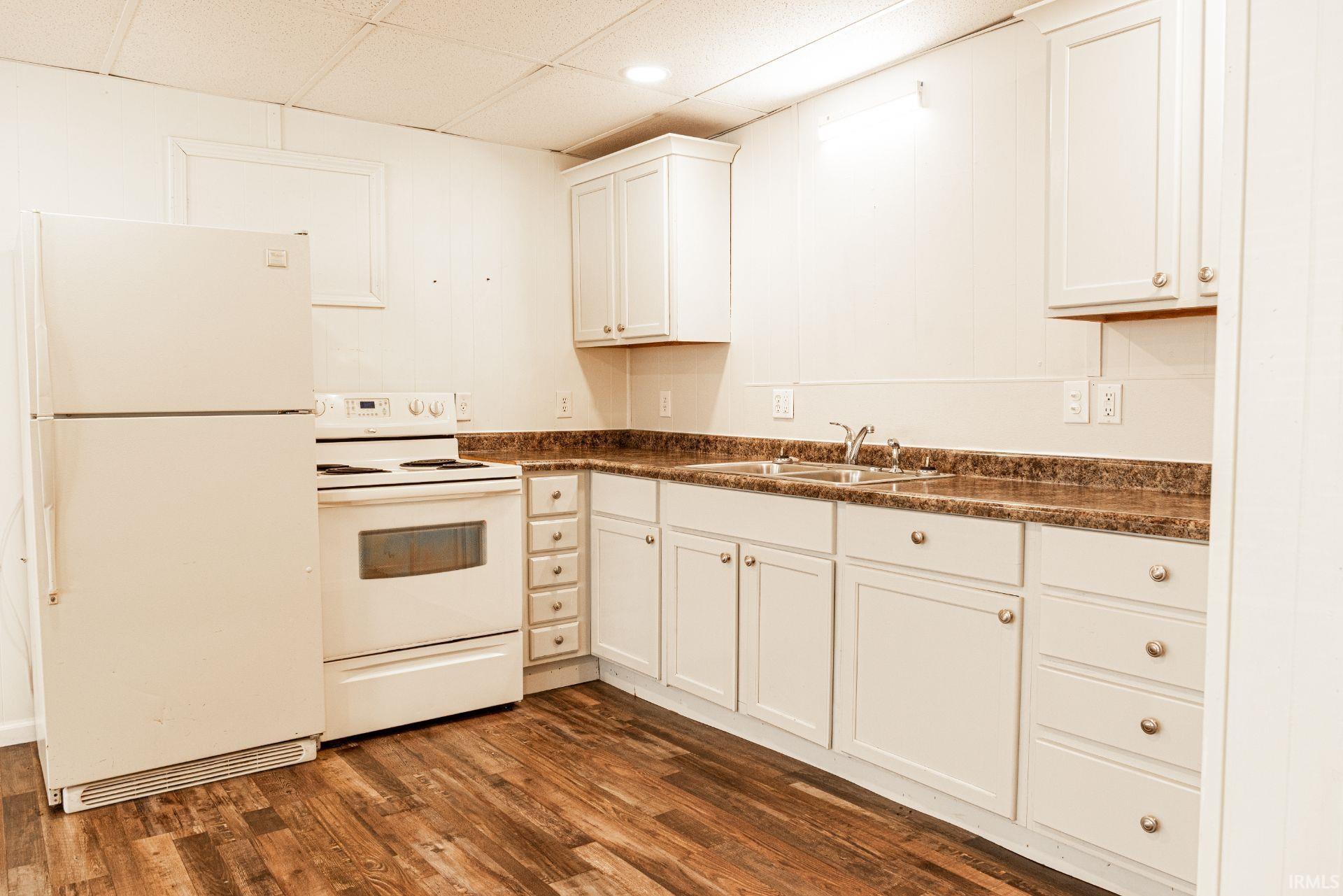 Kitchen featuring dark countertops, white appliances, dark wood-type flooring, a drop ceiling, and white cabinetry
