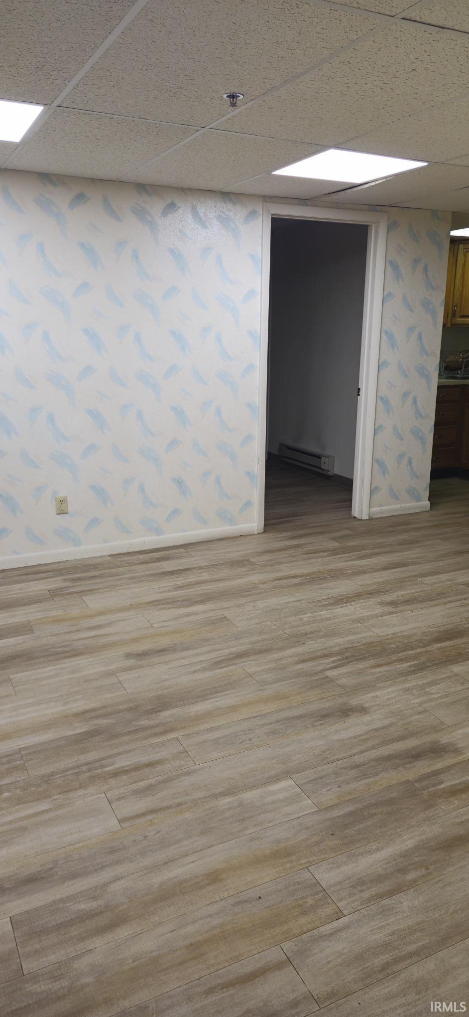 Finished below grade area featuring a drop ceiling, wallpapered walls, light wood-style floors, and a baseboard heating unit