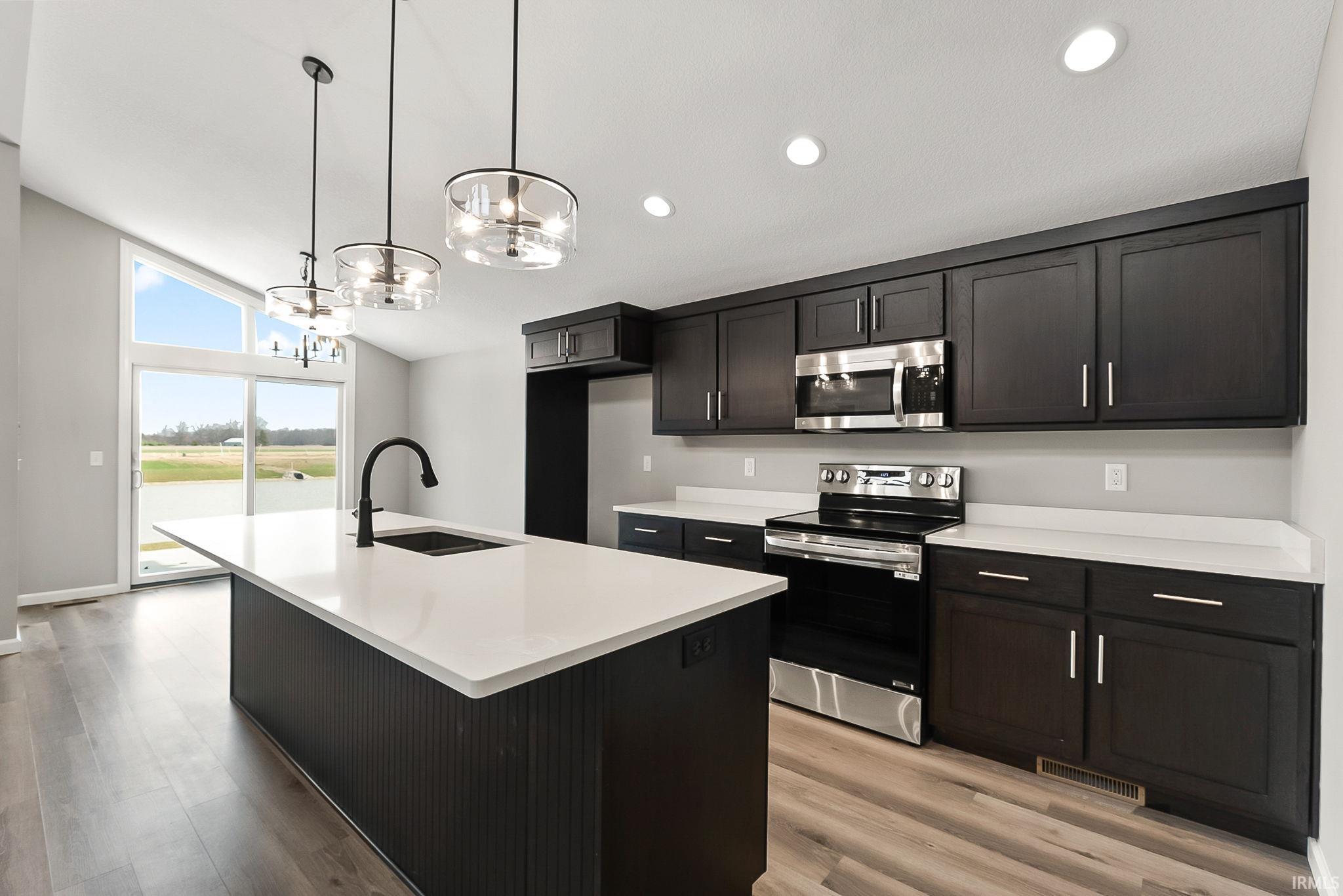 Kitchen featuring stainless steel appliances, a kitchen island with sink, light wood-style flooring, suspended lighting, and lofted ceiling