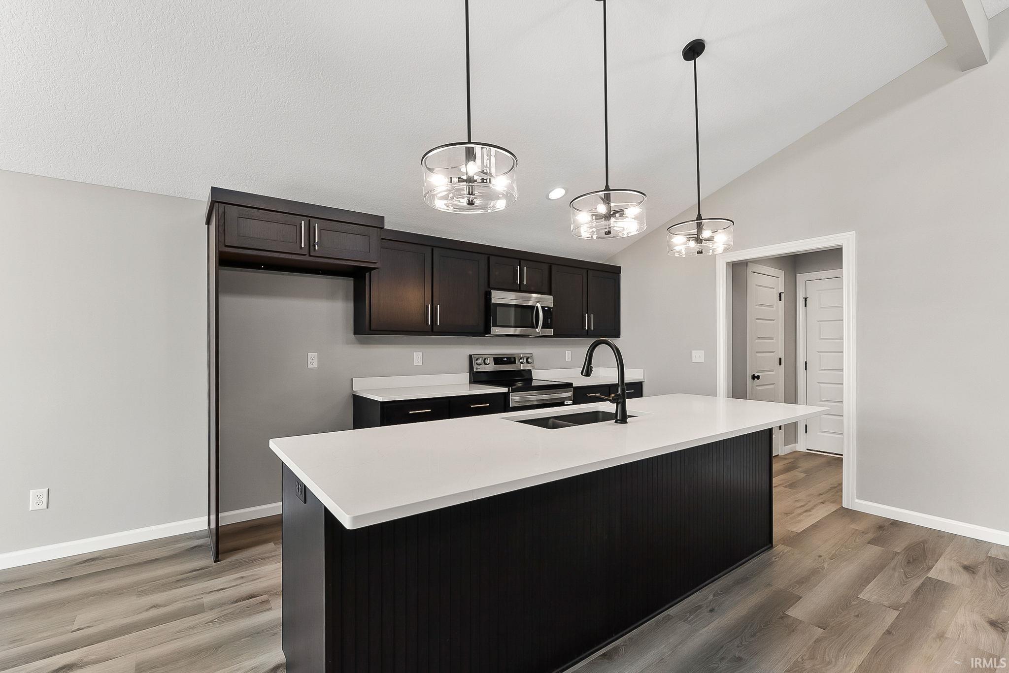 Kitchen with stainless steel appliances, hanging lights, a center island with sink, light wood-style floors, and lofted ceiling
