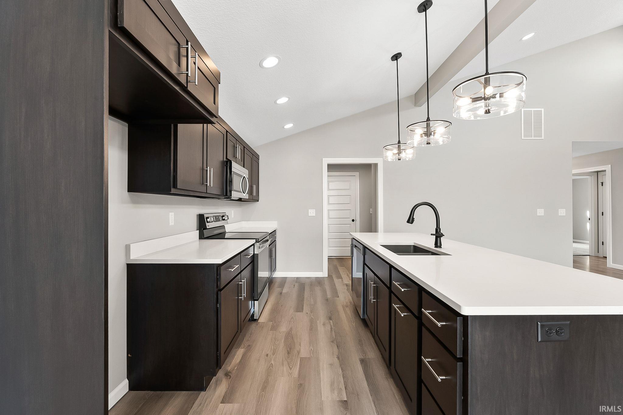 Kitchen featuring stainless steel appliances, a center island with sink, light wood-style floors, dark wood finish cabinets, and lofted ceiling with beams