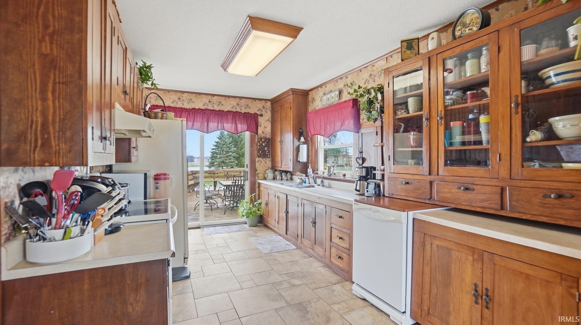 Kitchen with wood finish cabinets, glass insert cabinets, white dishwasher, wallpapered walls, and light countertops