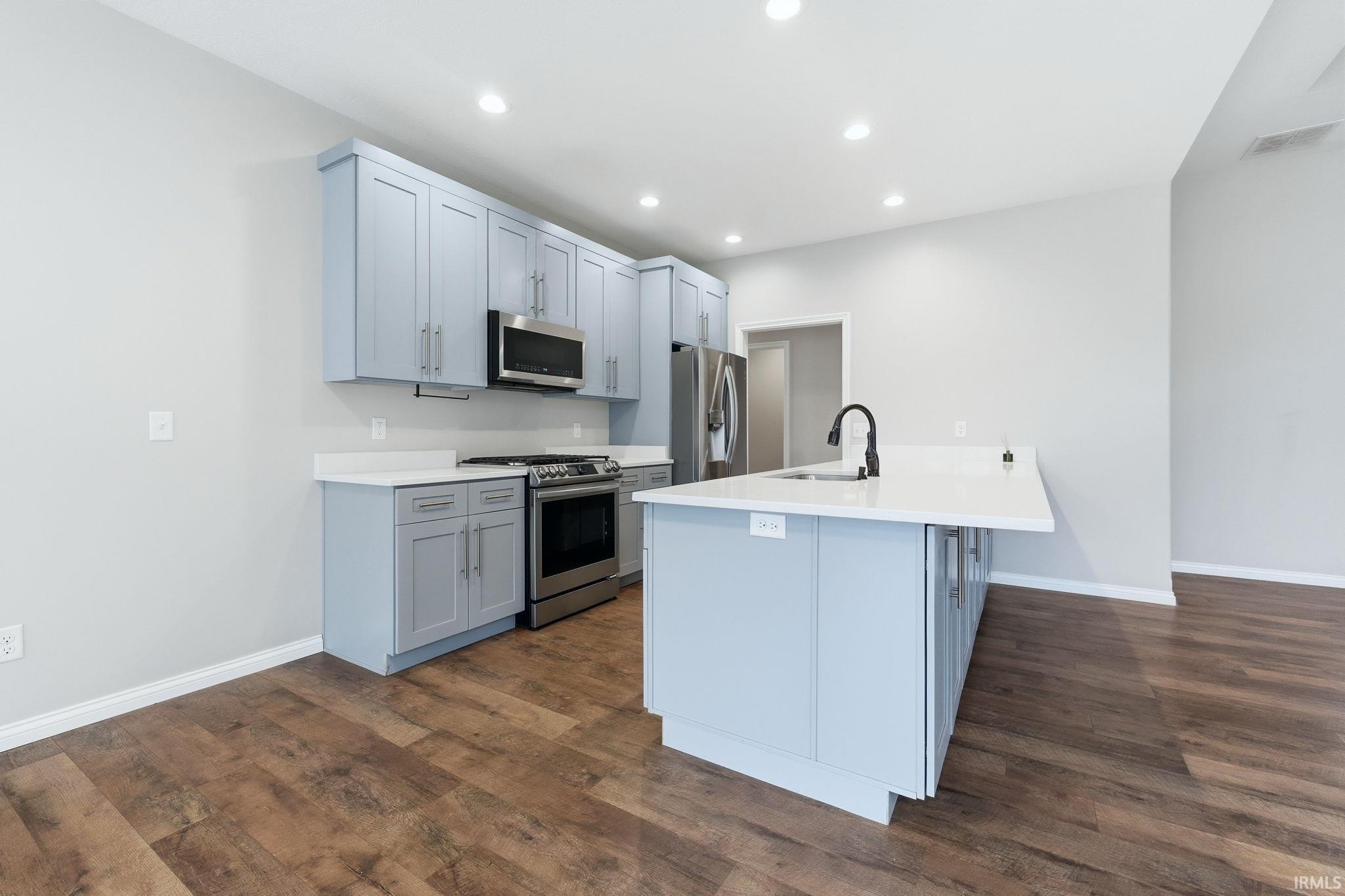 Kitchen with gray cabinets, stainless steel appliances, dark wood-style floors, recessed lighting, and an island with sink