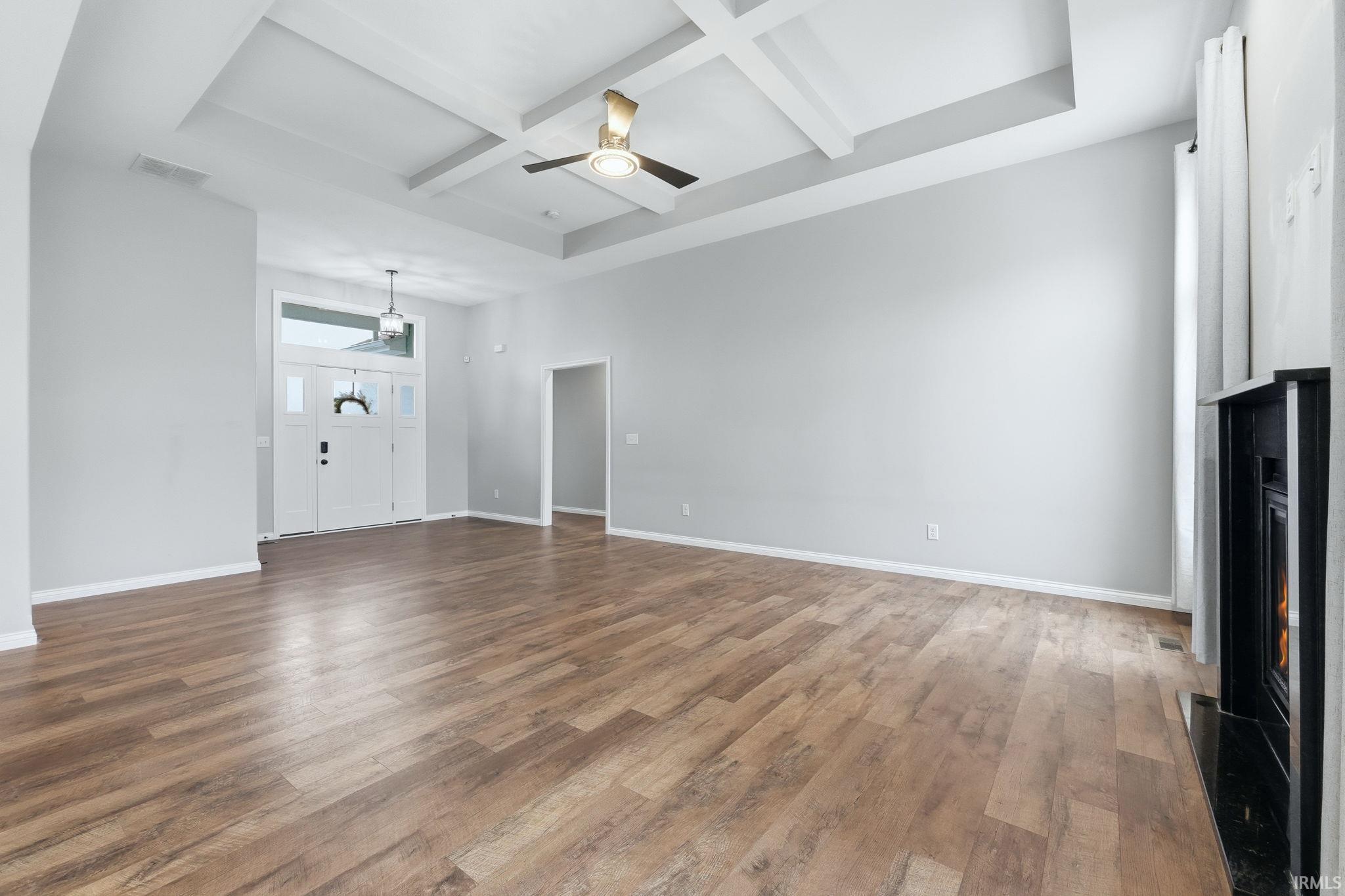 Unfurnished living room featuring coffered ceiling, ceiling fan, a warm lit fireplace, and dark wood-style floors