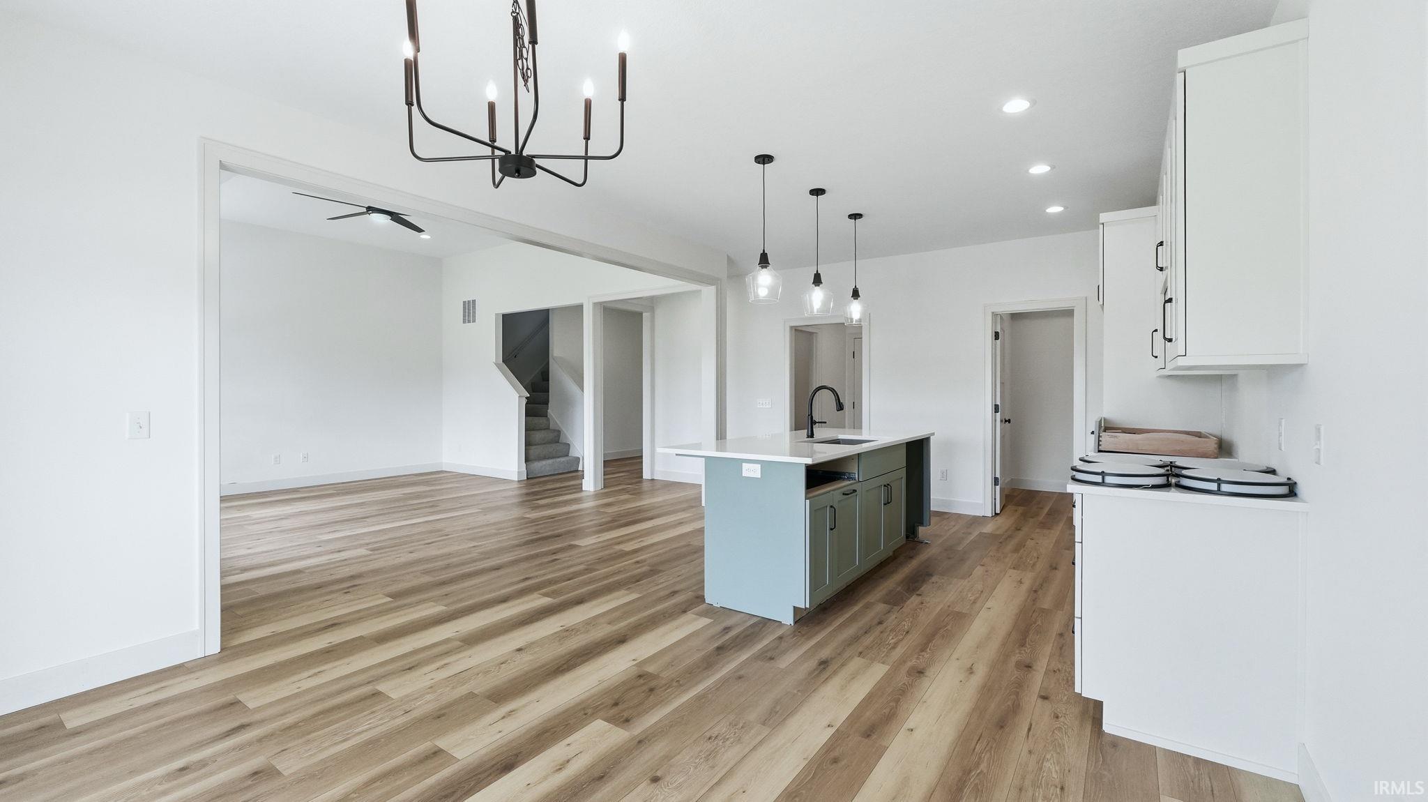 Expansive open-concept living area featuring wood-finish flooring, a modern kitchen island with pendant lighting, white cabinetry with black hardware, recessed lighting, and a contemporary black chandelier