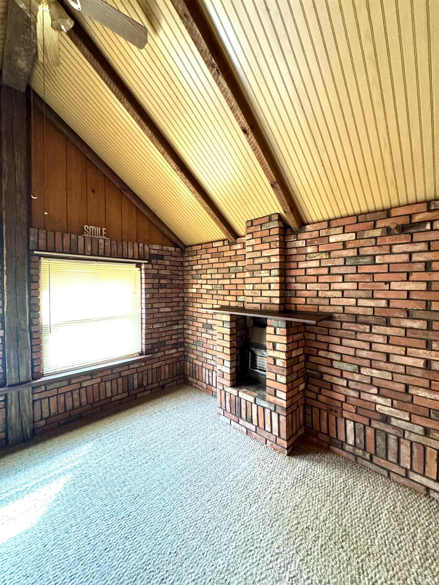 Unfurnished living room with lofted ceiling with beams, carpet floors, a brick fireplace, a ceiling fan, and brick wall