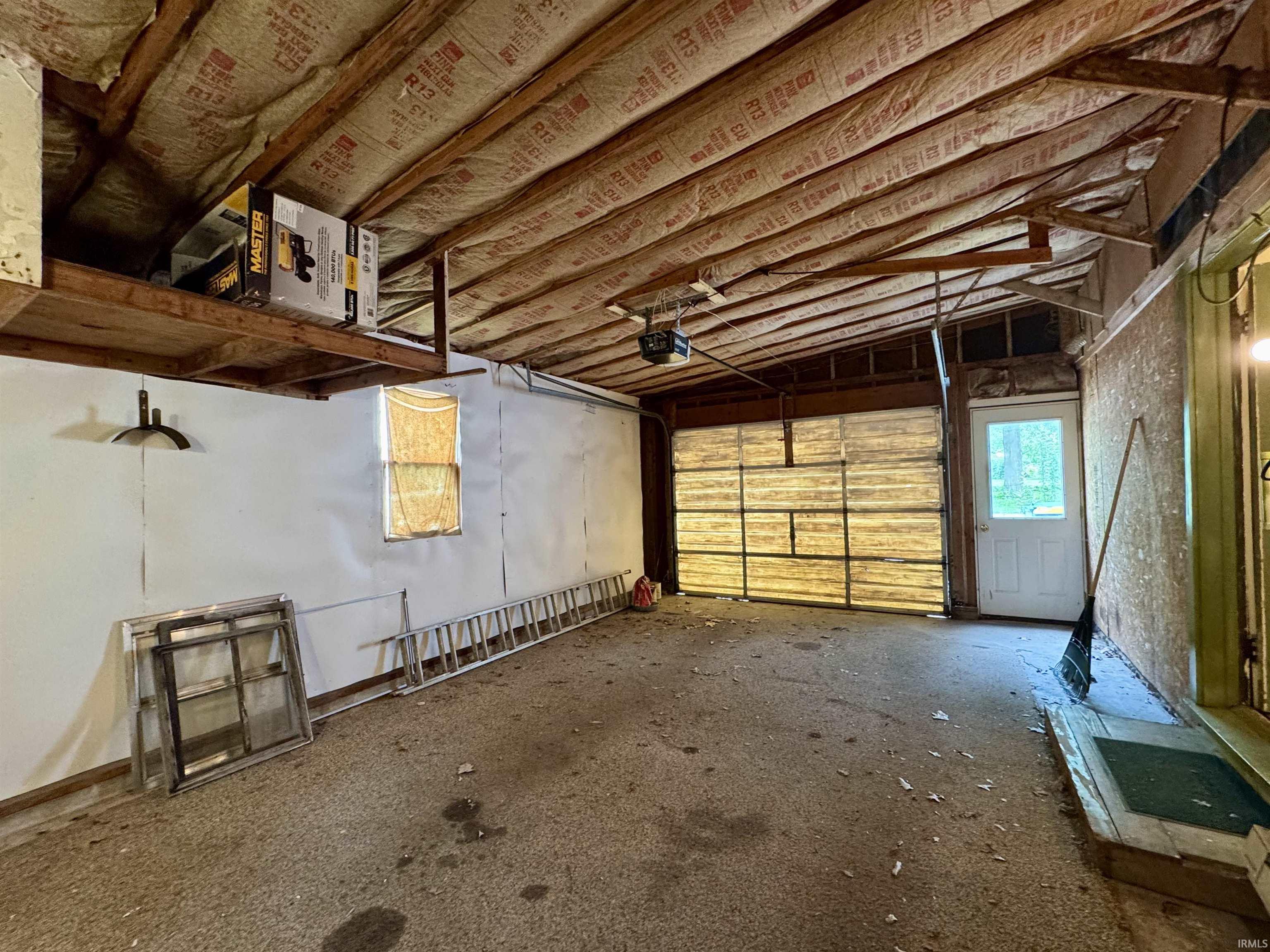 Spacious garage area featuring an exposed beam ceiling with insulation, a large garage door, a side entry door with glass paneling, a storage shelf, and concrete flooring