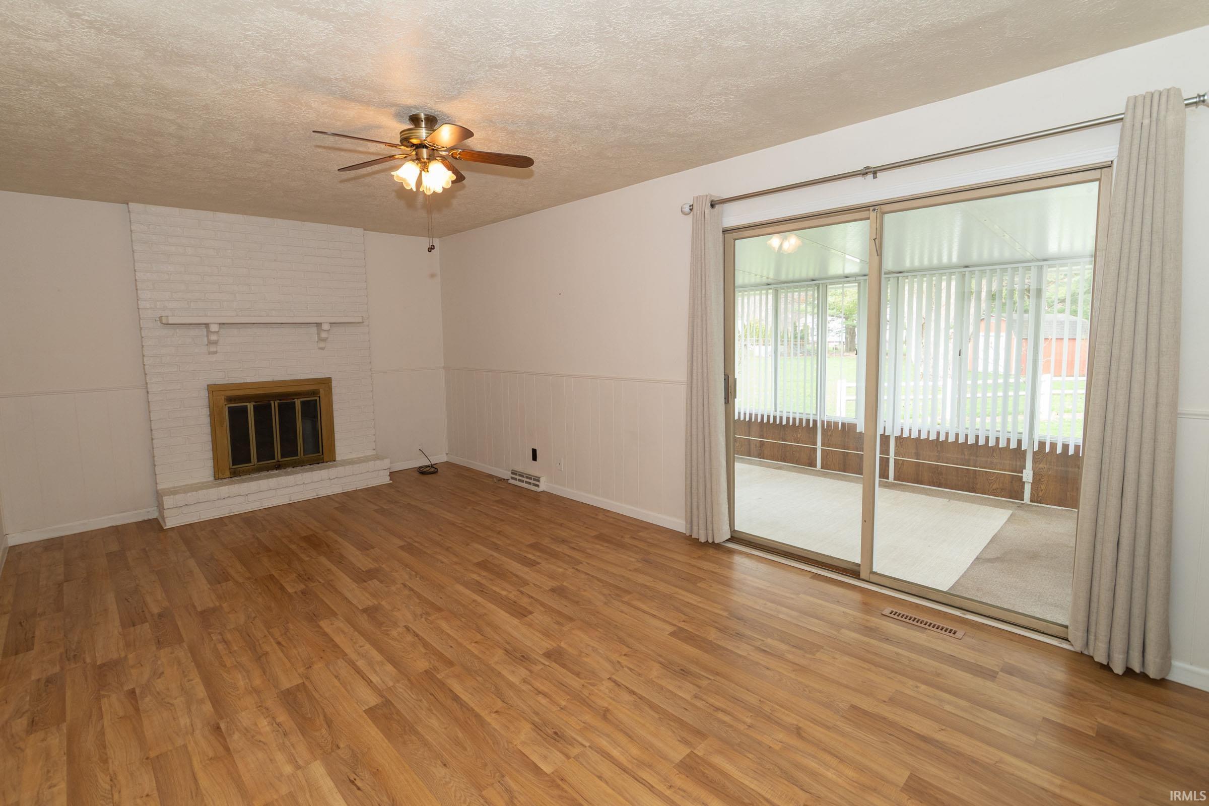 Unfurnished living room featuring a textured ceiling, a ceiling fan, light wood-style floors, a fireplace, and a wainscoted wall